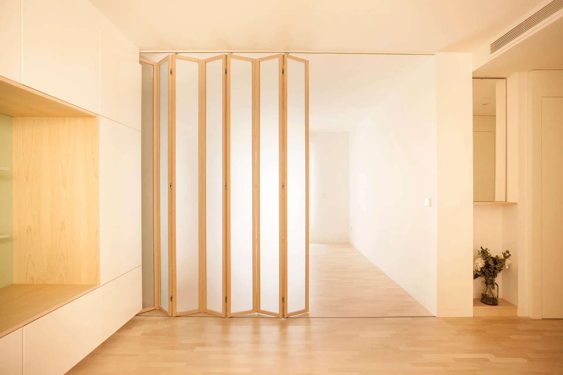 A minimalist room featuring a light wood folding screen with translucent panels, light wood flooring, and built-in white cabinetry with a wood niche.