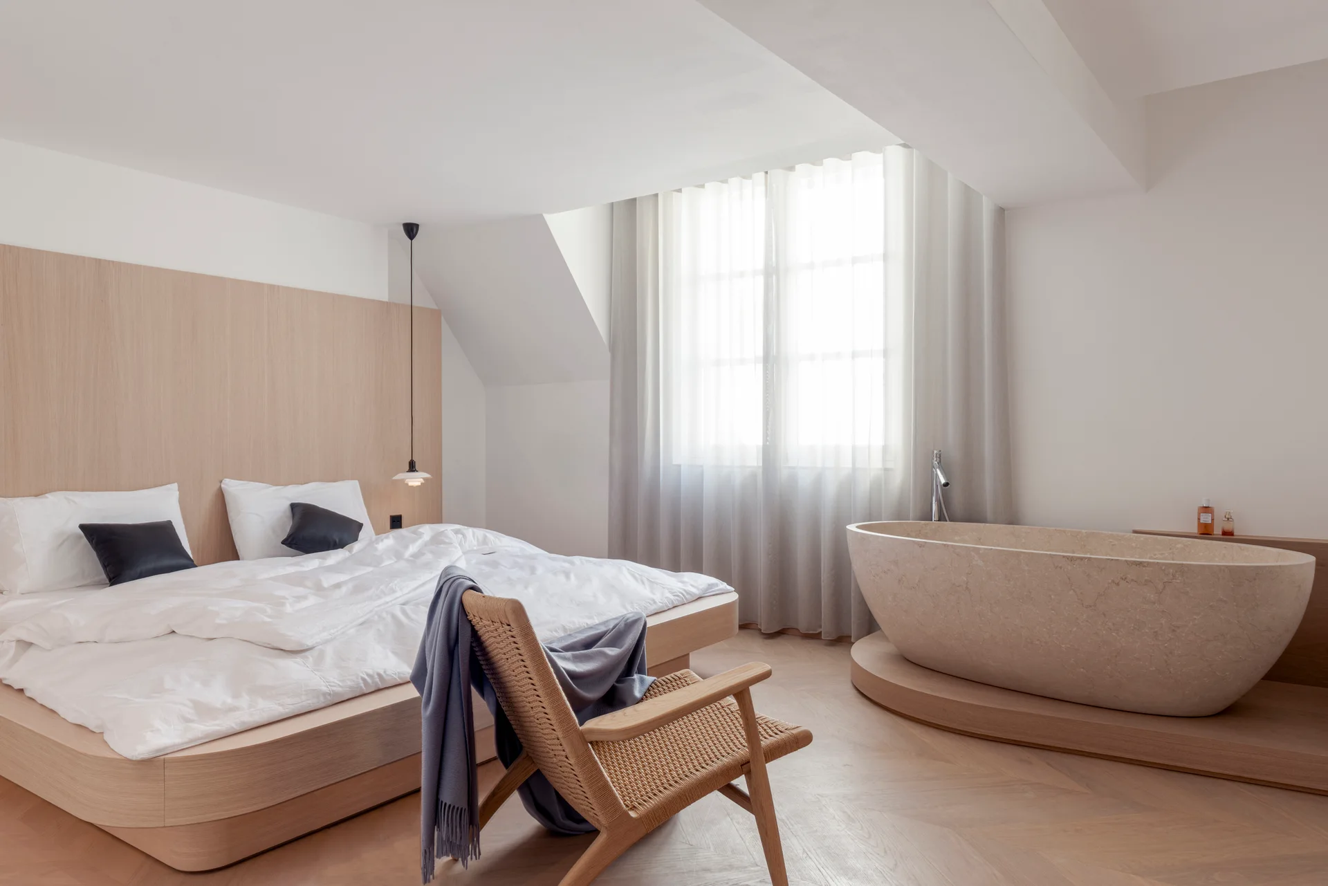 A minimalist bedroom features a wooden platform bed and a freestanding stone bathtub on a raised wood platform near a window with sheer curtains.