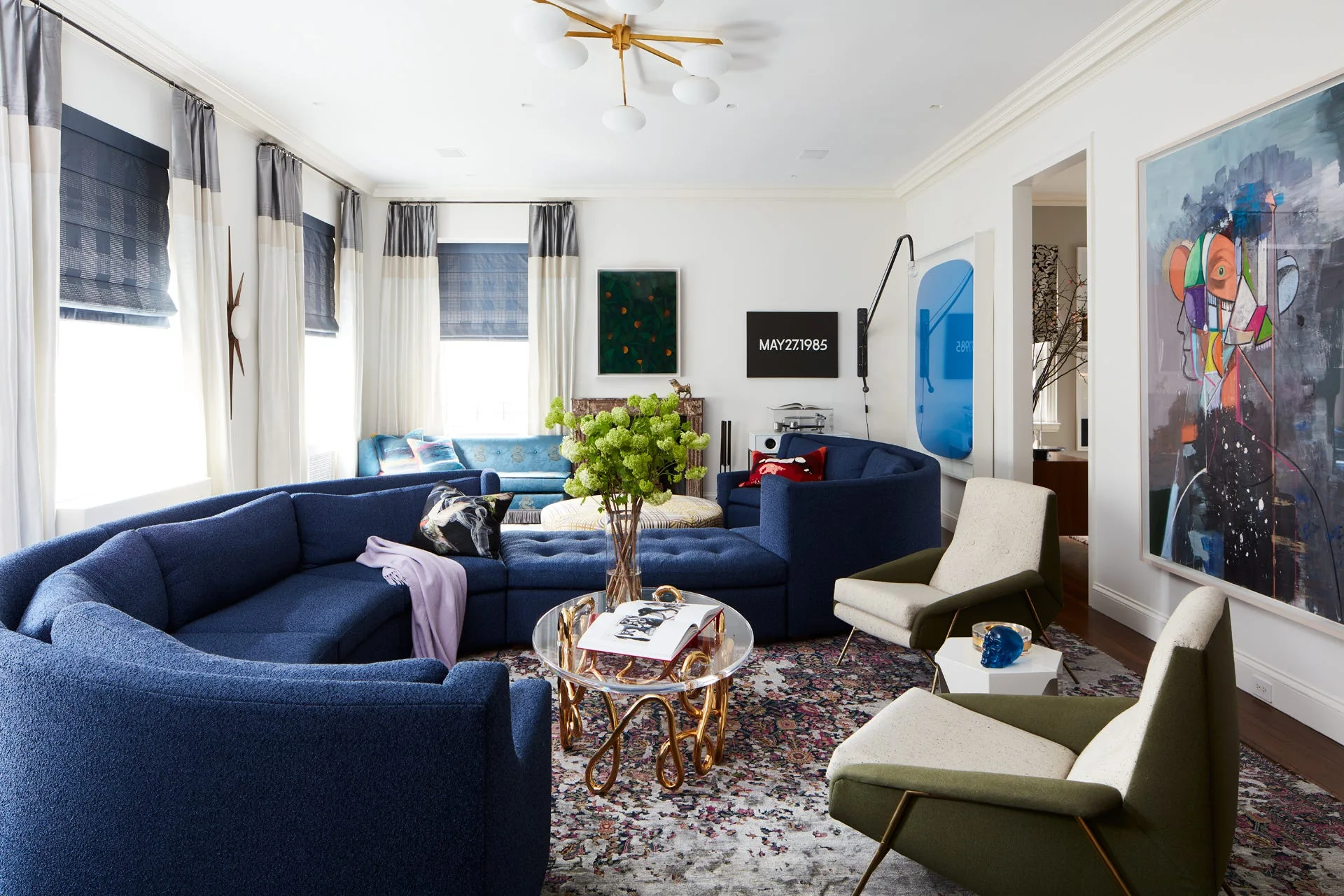 A contemporary living room features a large curved blue sectional sofa, two olive green armchairs, a patterned rug, and colorful modern wall art.