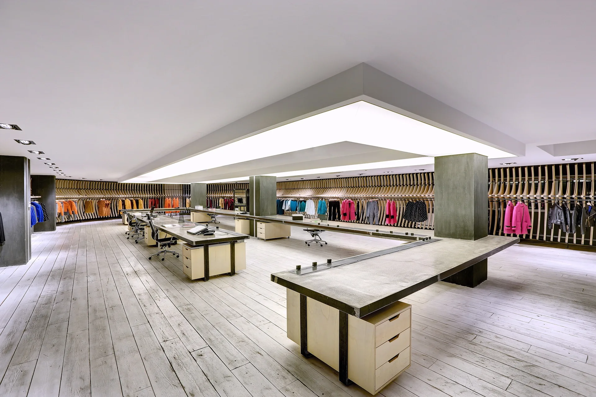 A wide commercial showroom featuring long custom wood workstations, concrete columns, and curved wood slat clothing racks on the walls.