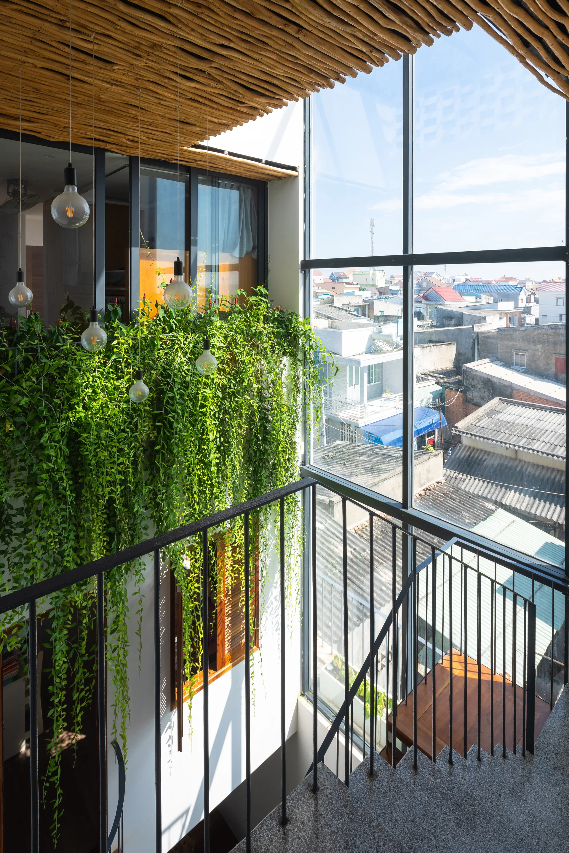 A concrete staircase with black metal railings features a wood stick ceiling, hanging lightbulbs, and cascading green plants beside a tall window.