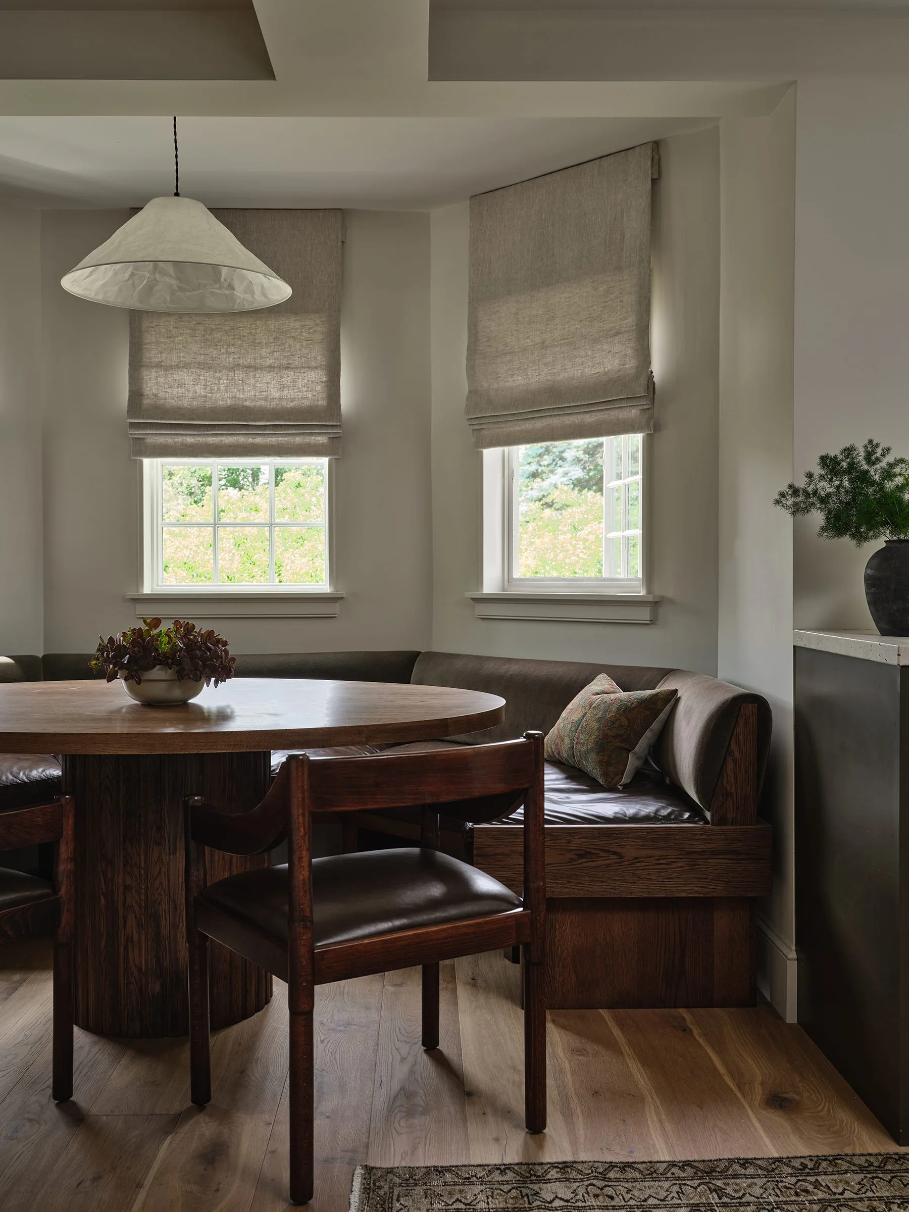 A corner dining nook features a built-in wooden banquette with leather cushions, a round pedestal table, and windows with linen roman shades.