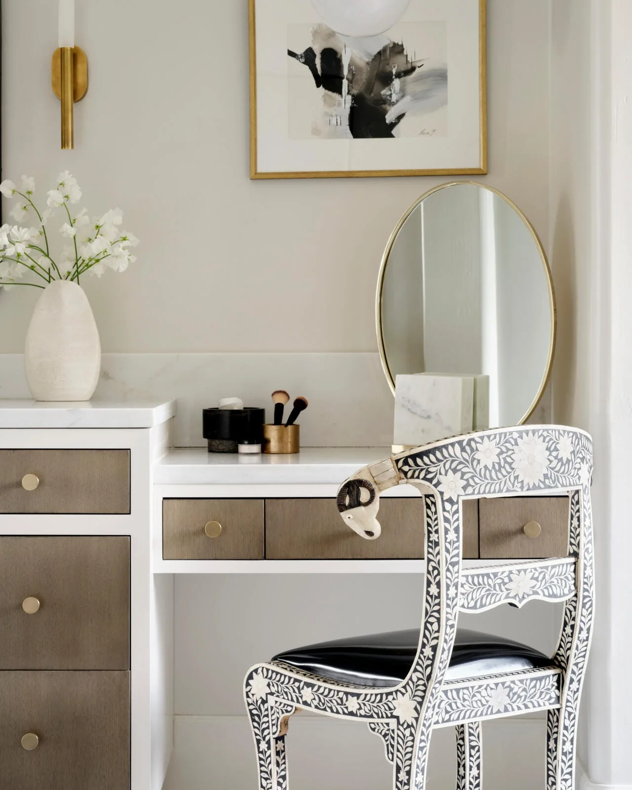 A white vanity with a marble countertop features gray drawers, an oval gold tabletop mirror, and an ornate floral inlaid chair.