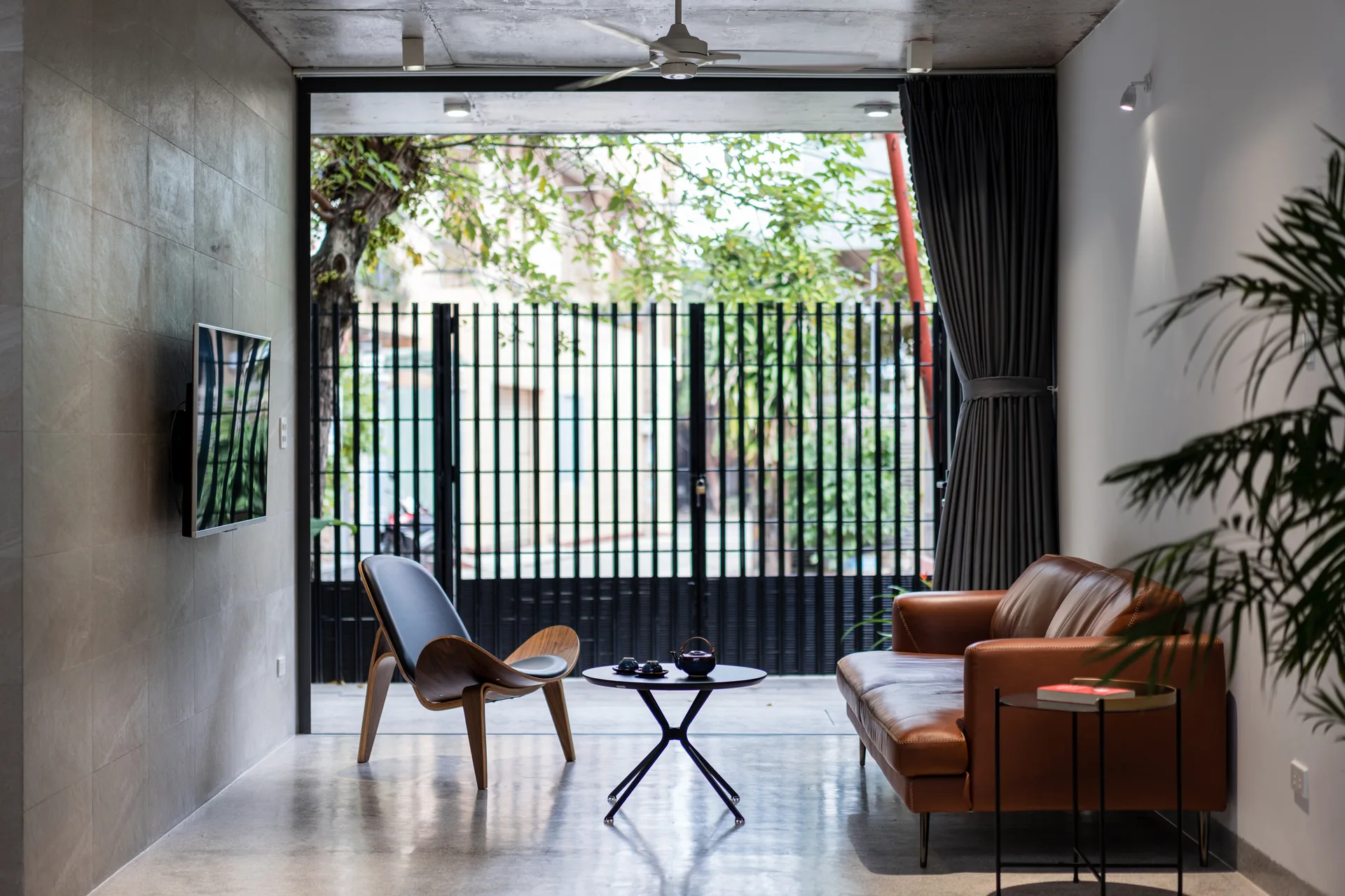 A modern living room features a brown leather sofa, a curved wood accent chair, and a concrete ceiling opening directly to a gated outdoor area.