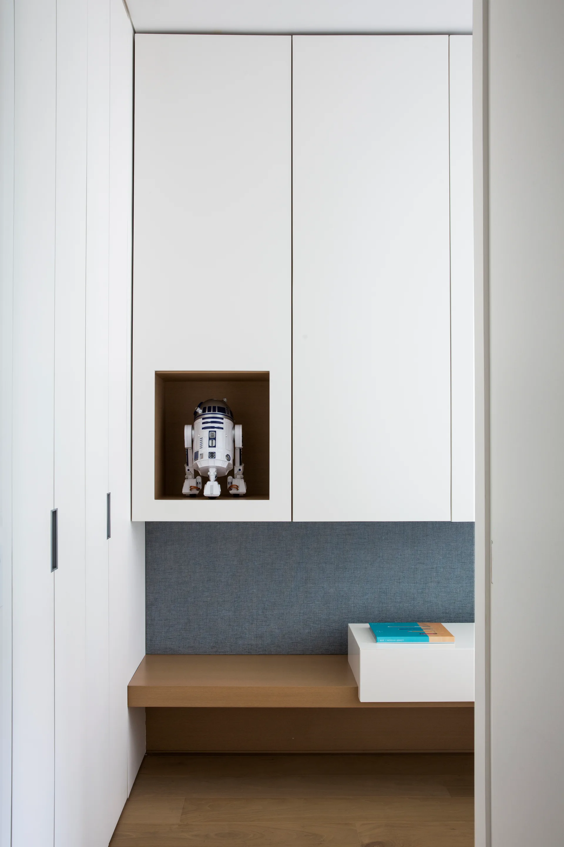 Modern white built-in storage cabinets feature a wooden bench below a grey fabric-covered wall panel and a small square niche holding a toy robot.