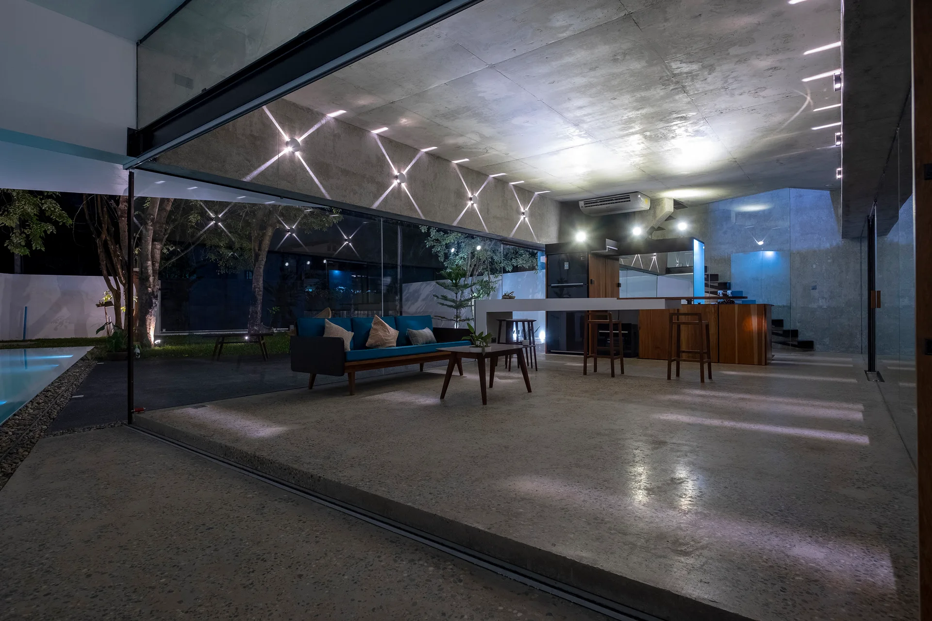 A modern living room with an exposed concrete ceiling, a blue sofa, and large sliding glass walls that open to an outdoor pool area.