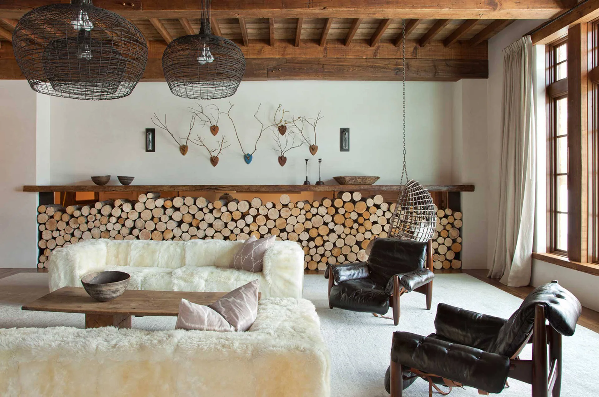 A rustic living room features plush cream sheepskin sofas, dark leather armchairs, exposed ceiling beams, and a wall filled with stacked logs.