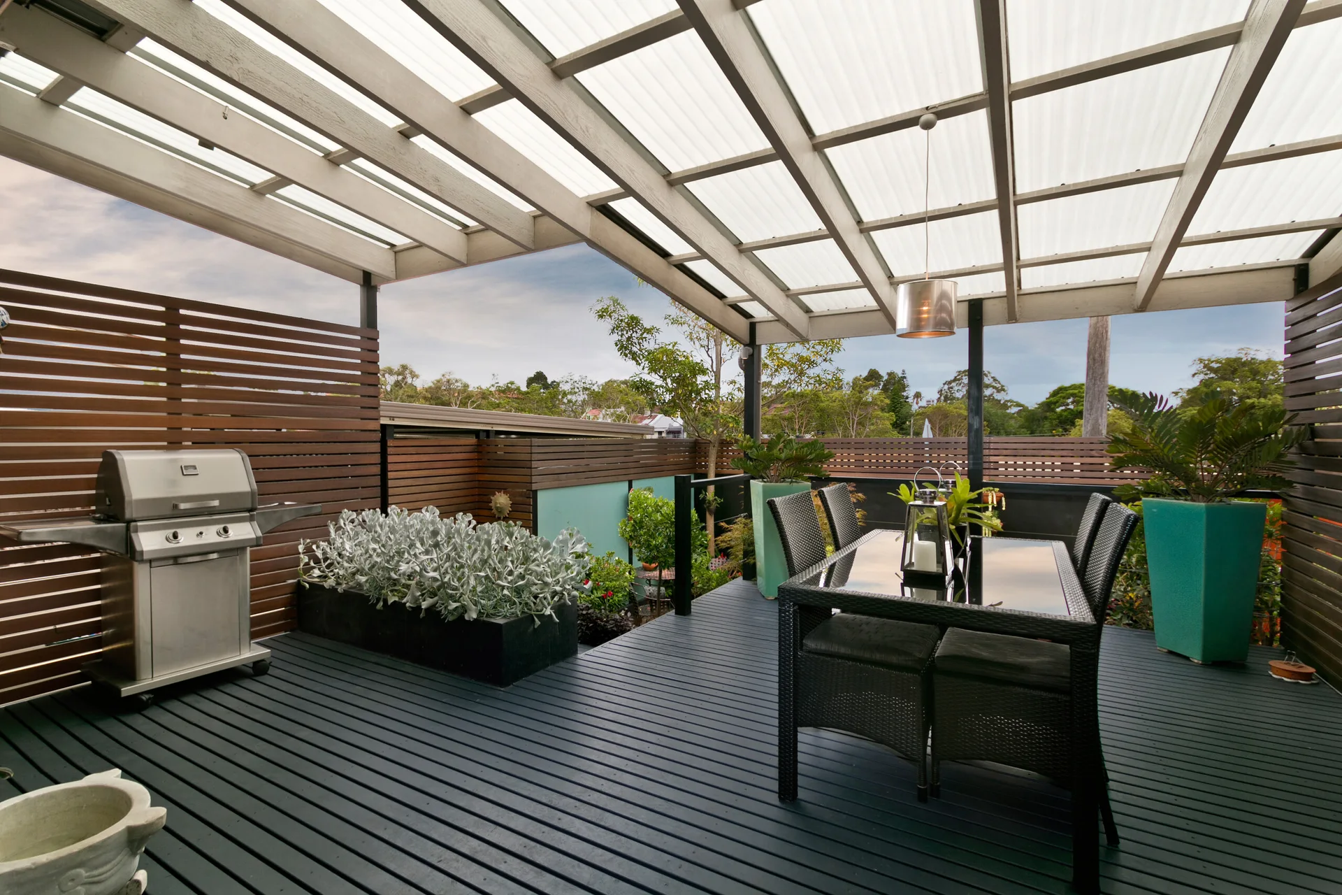 A covered outdoor deck features a black dining set under a slatted pergola, a stainless steel grill, and tall wood privacy screens.