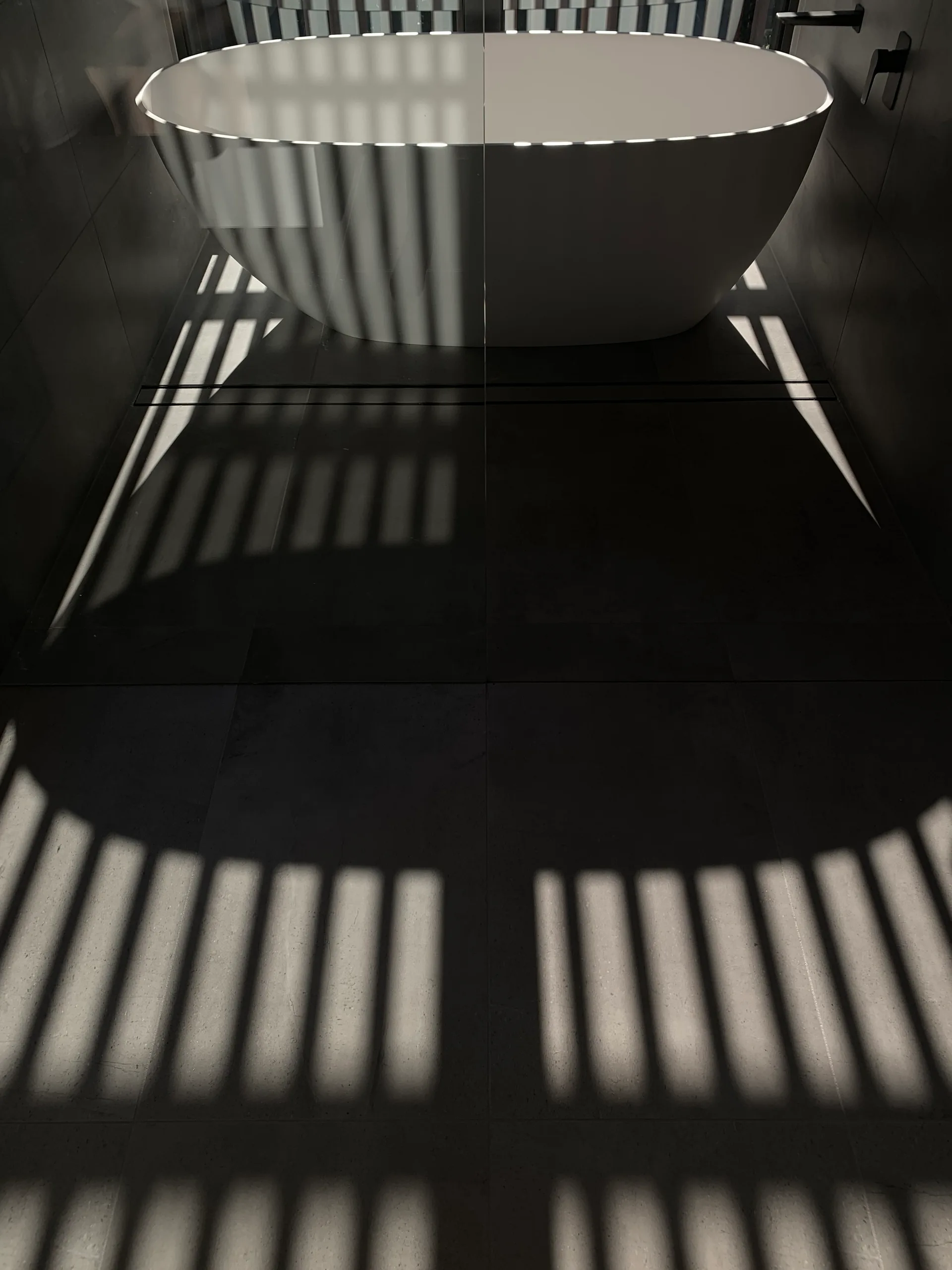 A white freestanding bathtub is set within a dark charcoal tiled bathroom with linear shadows cast across the floor.