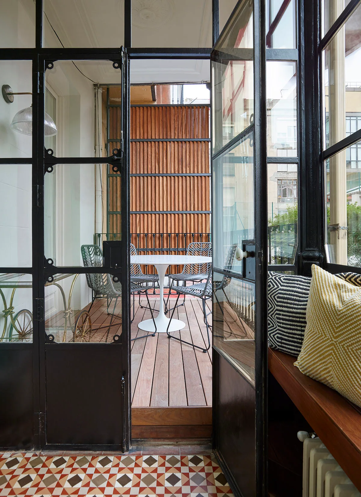 A view through black steel glass doors to a narrow wooden terrace featuring a round white pedestal table, wire chairs, and a vertical wood privacy screen.