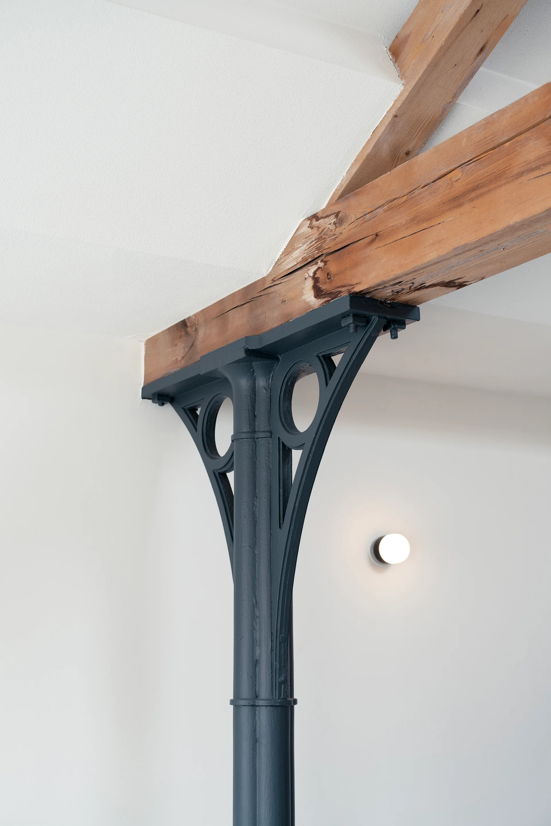 A dark charcoal painted cast iron column with decorative brackets supports an exposed natural wood ceiling beam against a white wall.