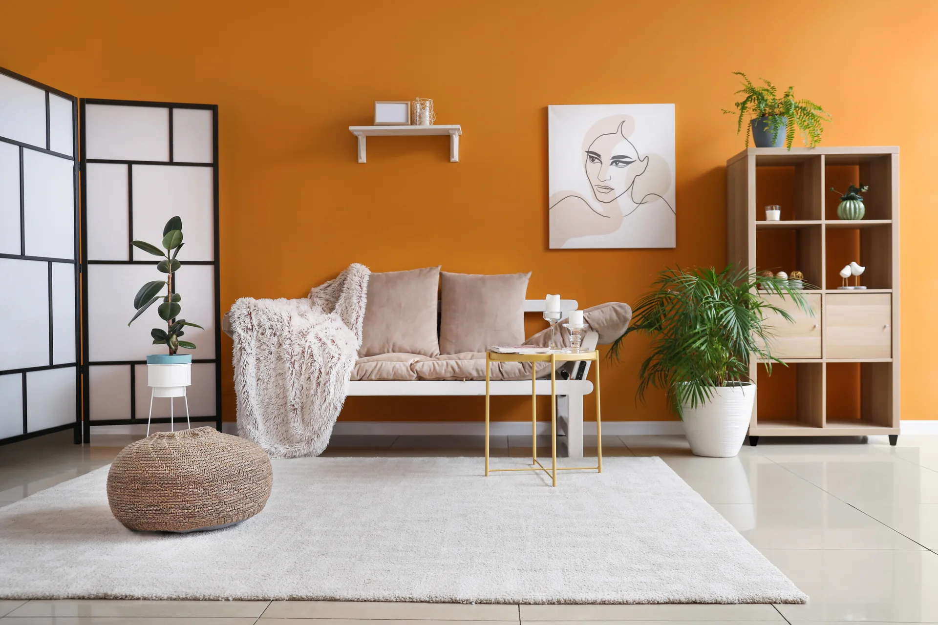 A modern living room features a bench-style sofa in front of a bright orange wall, a white rug, a woven pouffe, and a wooden bookcase.