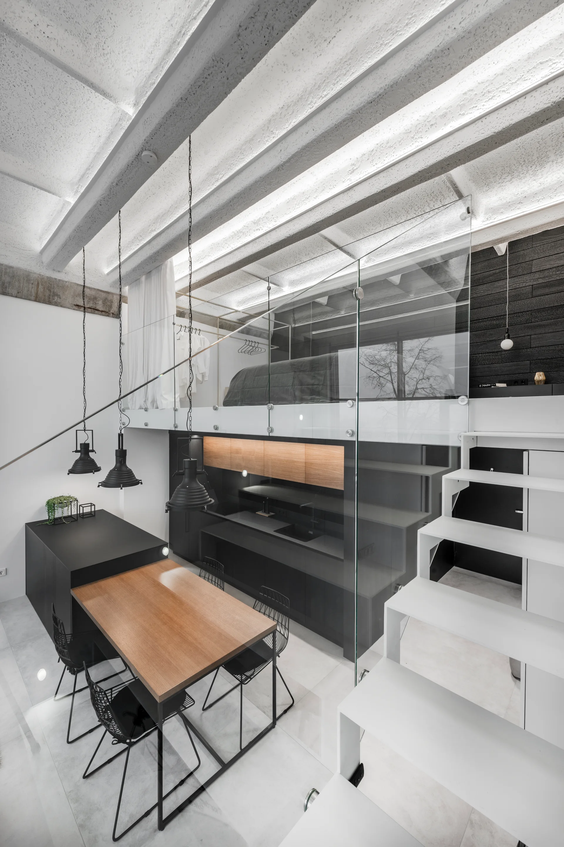 A modern loft interior featuring a white staircase, matte black kitchen, wood-topped dining table, and a glass-walled mezzanine bedroom.