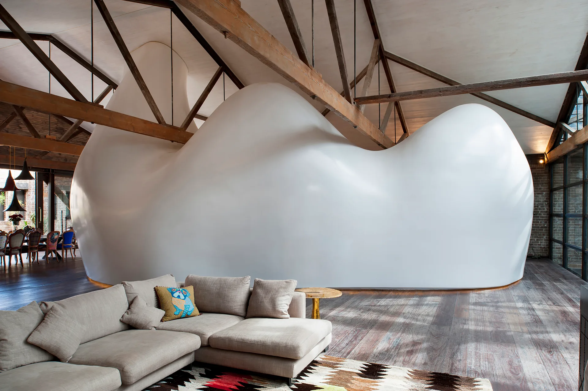 A spacious loft living room featuring a large, curved white sculptural partition wall beneath an exposed wooden truss ceiling.