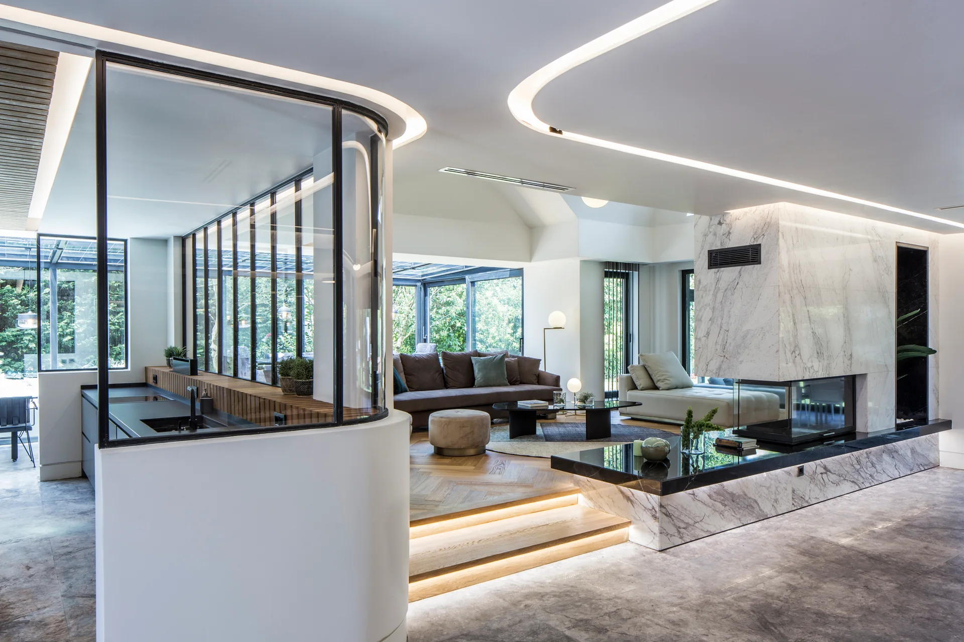 A modern sunken living room featuring a marble-clad fireplace, herringbone wood floors, a curved glass partition, and integrated ceiling cove lighting.