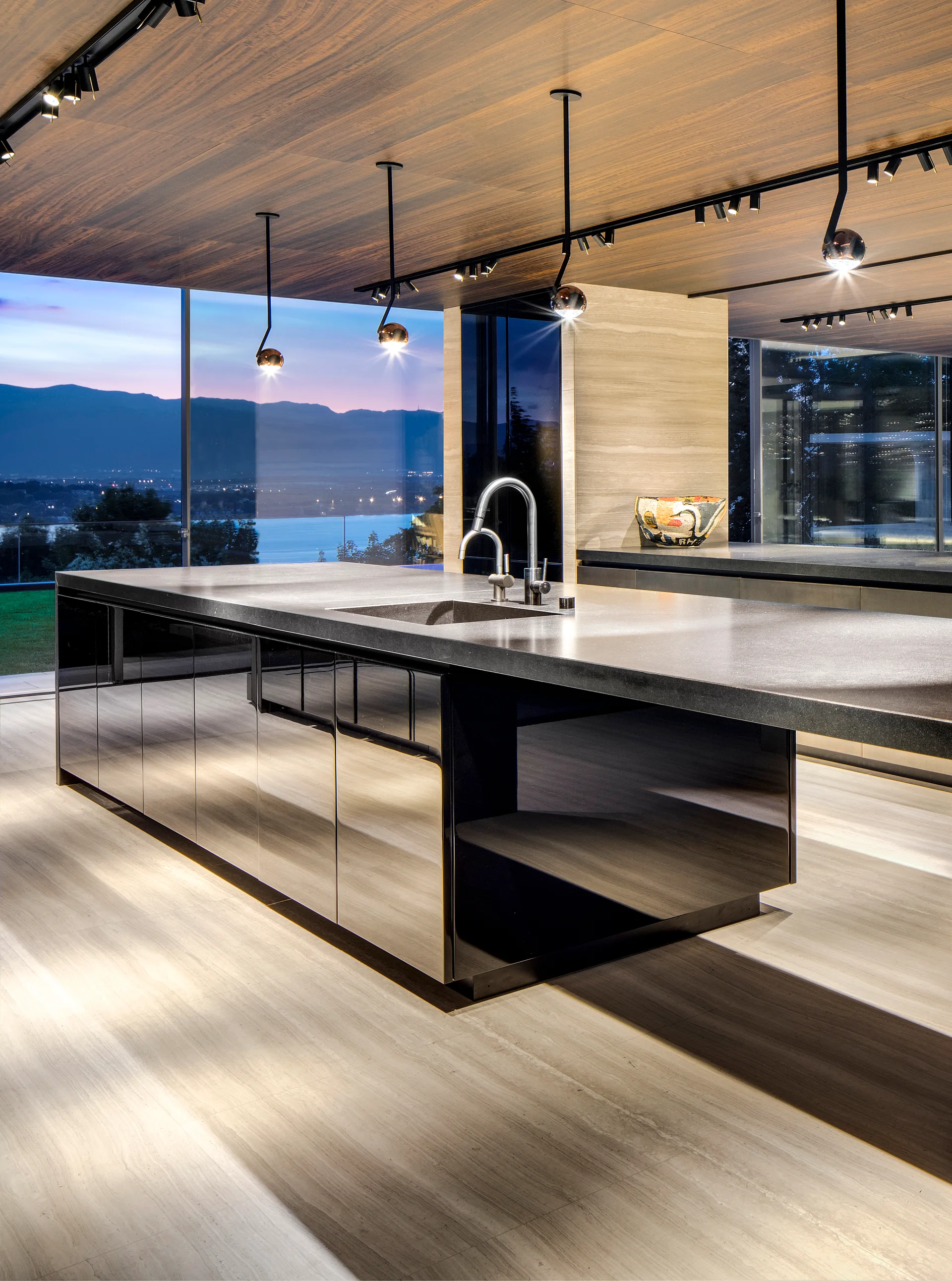 Minimalist kitchen featuring a glossy black island under a wood-paneled ceiling, with large windows showing a twilight mountain landscape.