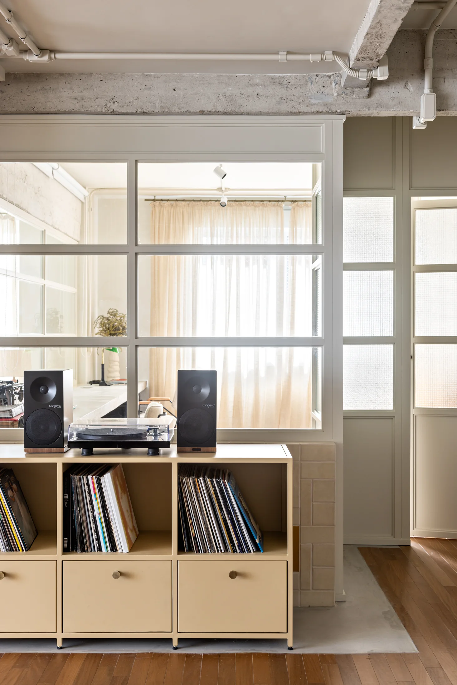A tan modular vinyl storage console with speakers and a turntable sits in front of a white-framed glass partition wall under a concrete ceiling.