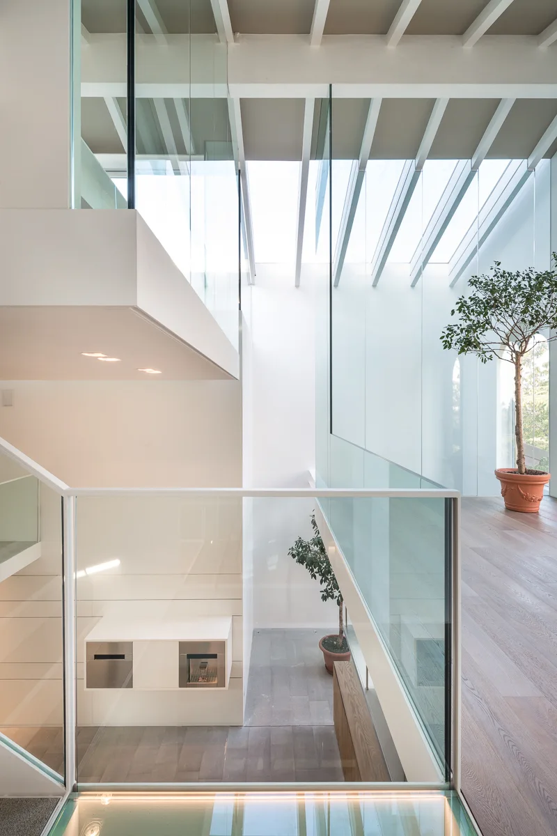 A multi-level minimalist interior features a modern staircase with glass balustrades, light wood flooring, white walls, and a large skylight.