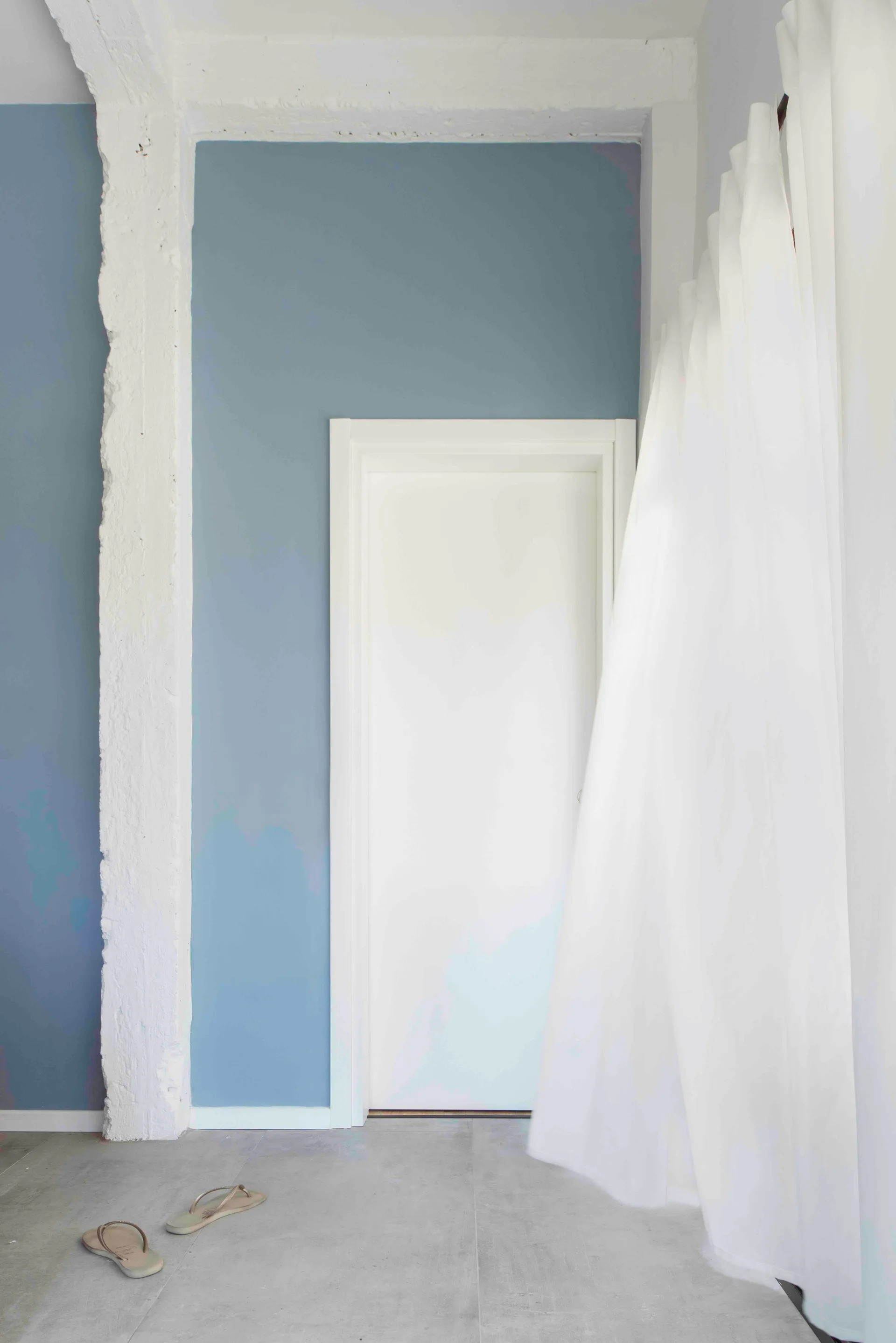 A minimalist interior featuring a white door on a blue-grey wall, situated between a rough textured column and sheer white curtains.