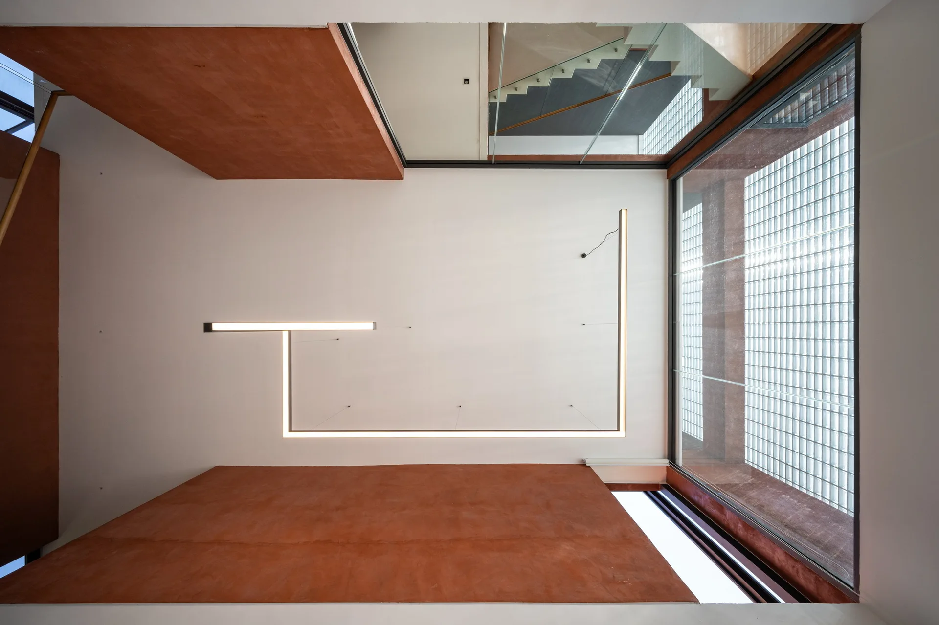 A top-down view of a modern stairwell featuring terracotta plaster walls, geometric linear LED lighting, and a glass block wall.