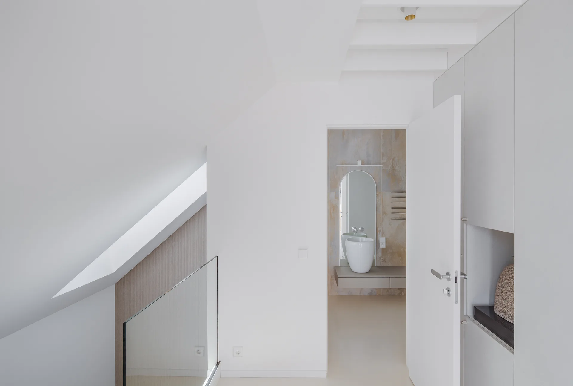 A view through a white doorway reveals a minimalist bathroom featuring an arched mirror, a bowl sink on a floating vanity, and a textured wall.