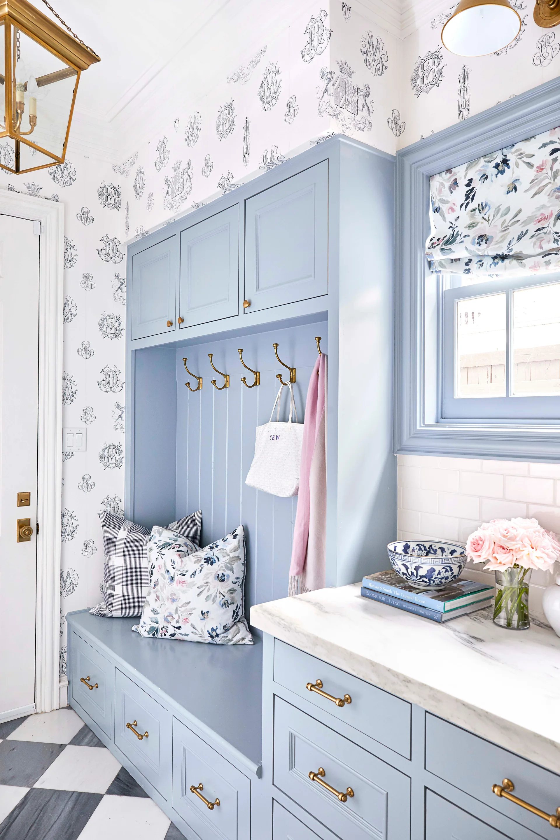 A light blue built-in mudroom bench features brass hooks and drawers, set against monogrammed wallpaper and checkered tile flooring.