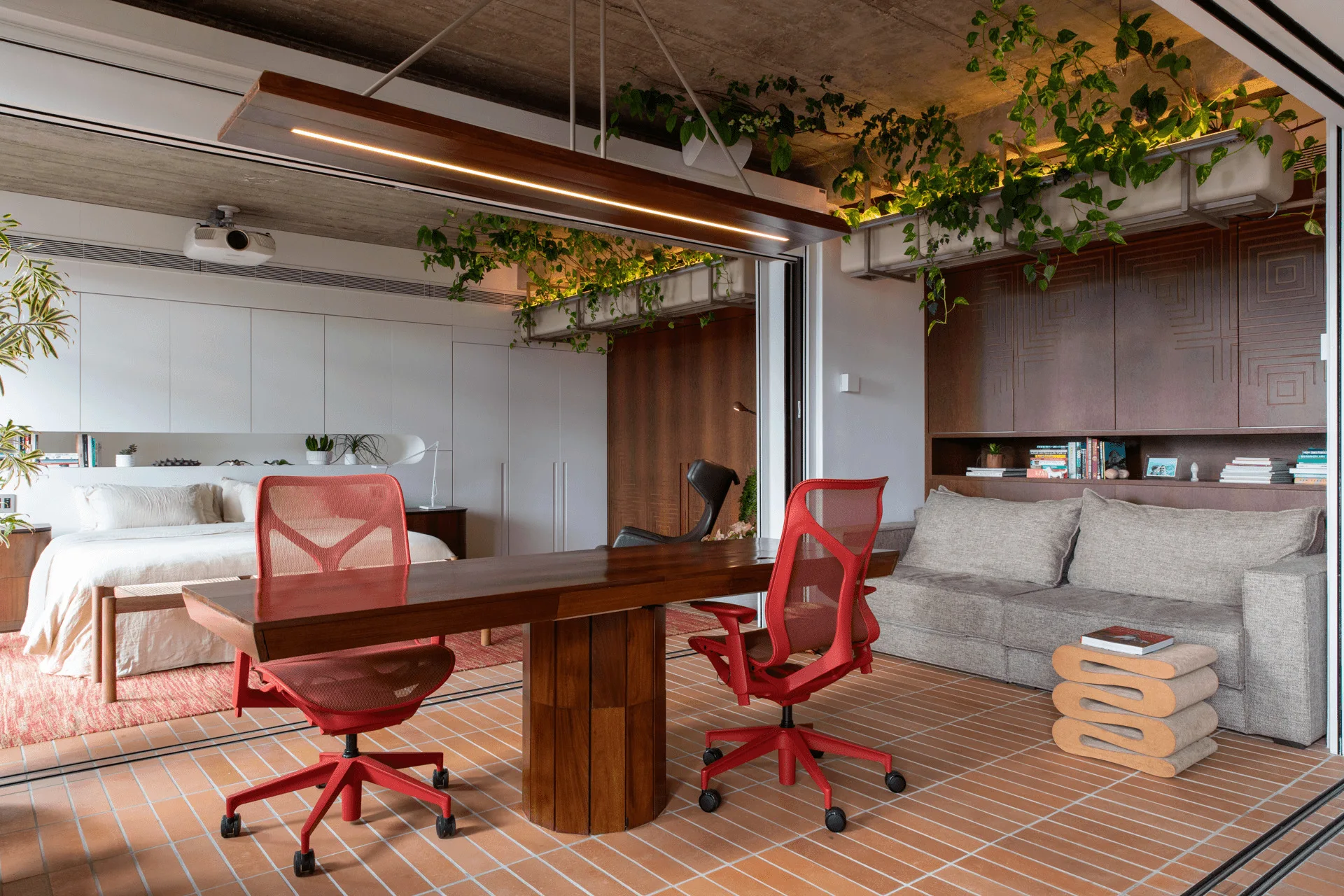 A contemporary studio interior featuring a wooden desk with two red office chairs, terracotta tiled floors, a grey sofa, and integrated ceiling plants.