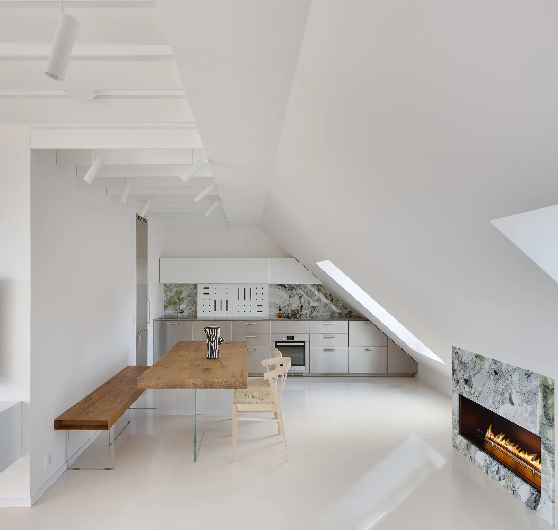 A minimalist attic kitchen with stainless steel cabinets, a wooden dining table with glass legs, and a built-in green marble fireplace.