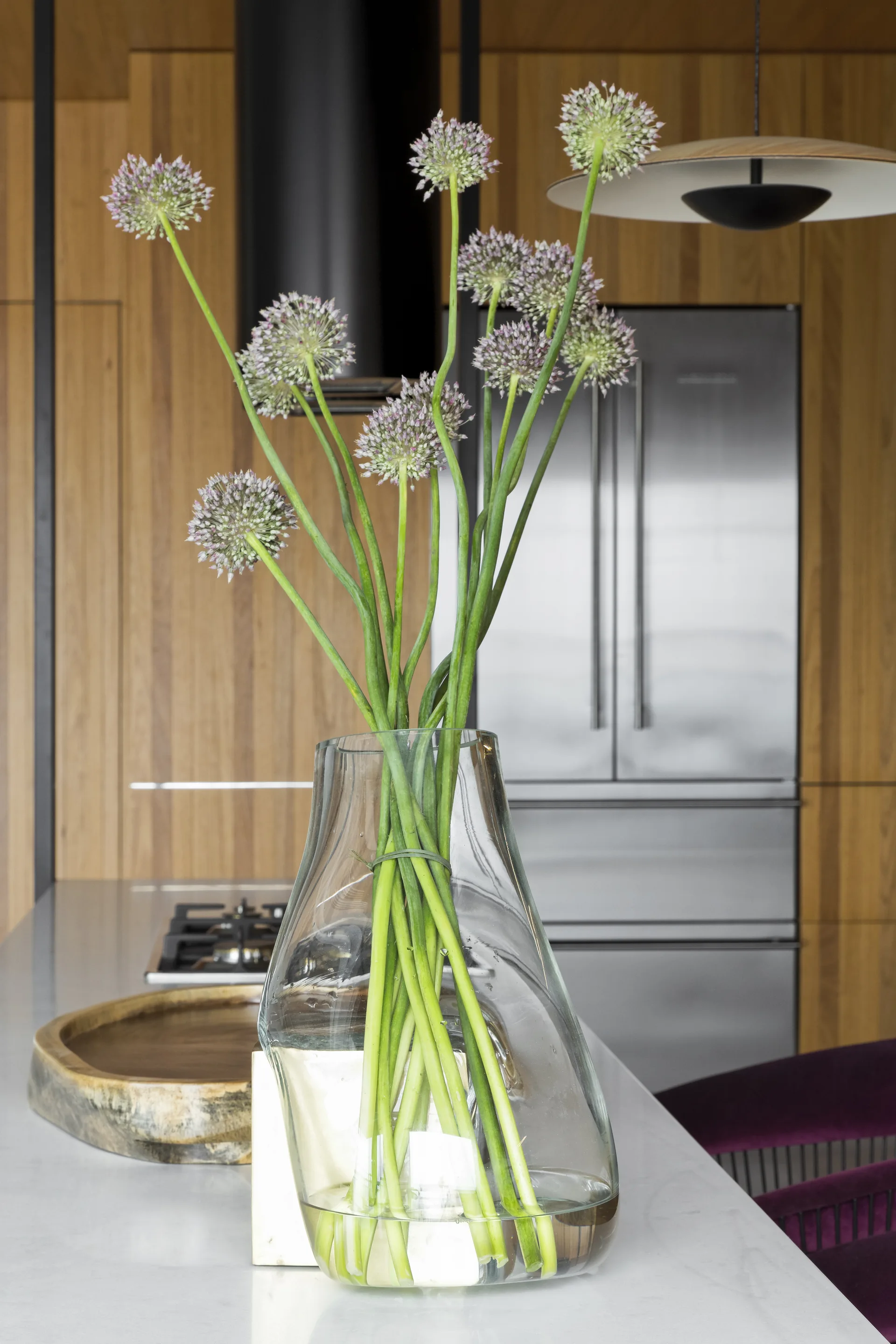 A tall glass vase filled with purple allium flowers sits on a white kitchen island in front of vertical-grain wood cabinets.