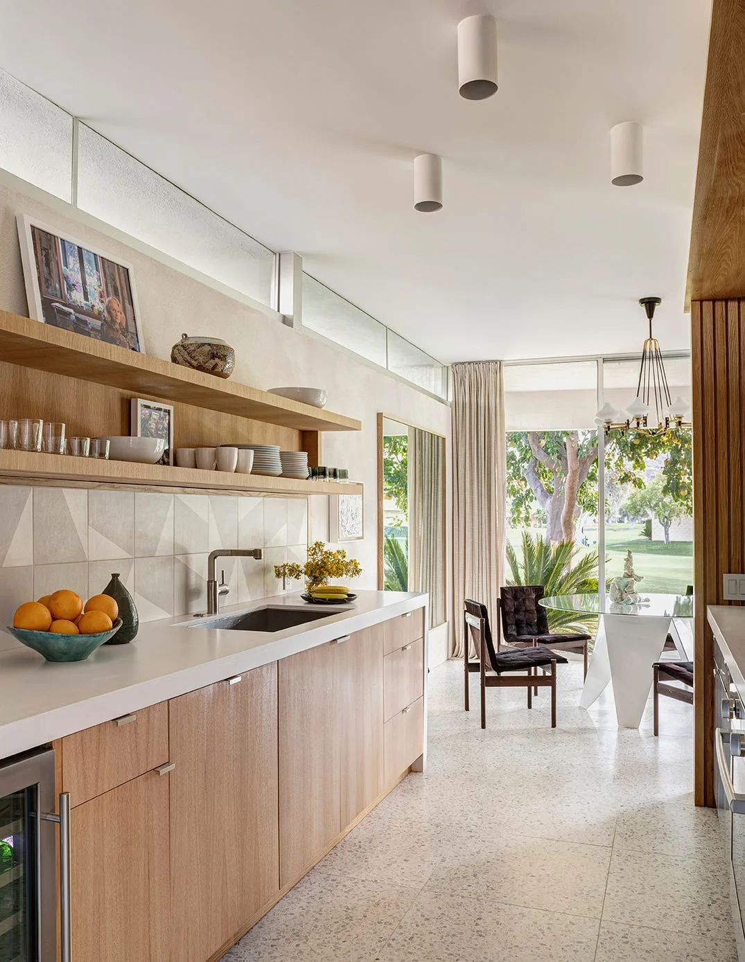 A modern kitchen featuring light wood cabinets, white countertops, floating shelves, a geometric tiled backsplash, and a dining table near a window.
