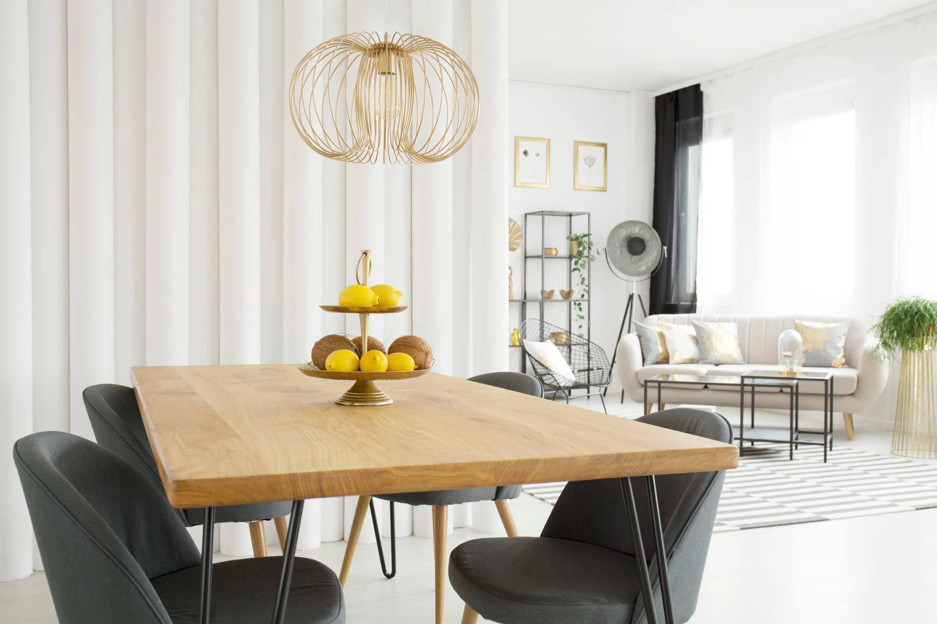 A wooden dining table with grey chairs sits in front of a white fabric divider, with a gold wire pendant light hanging above.