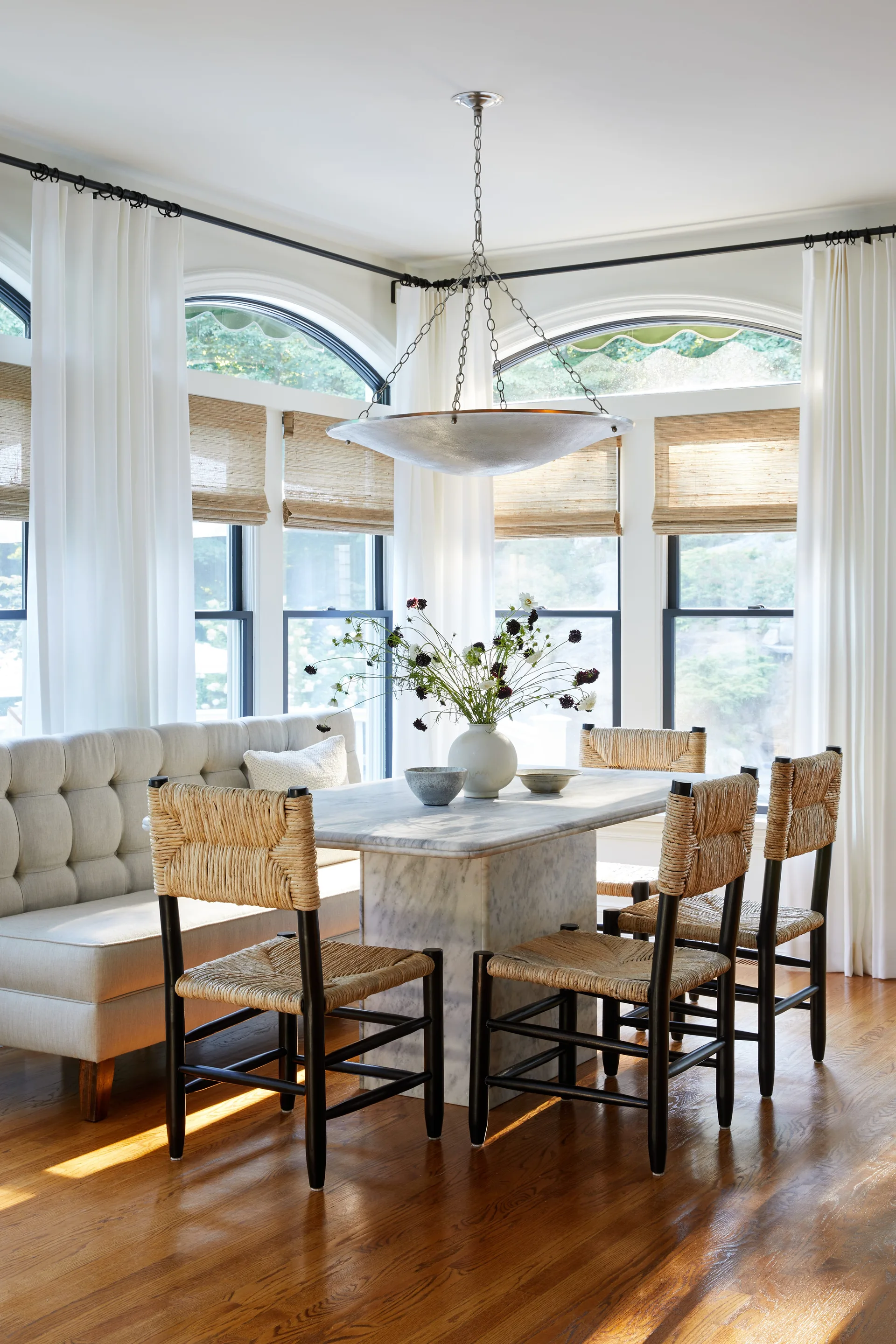 A sunlit breakfast nook features a marble pedestal table, a tufted cream banquette, black woven chairs, and arched windows.
