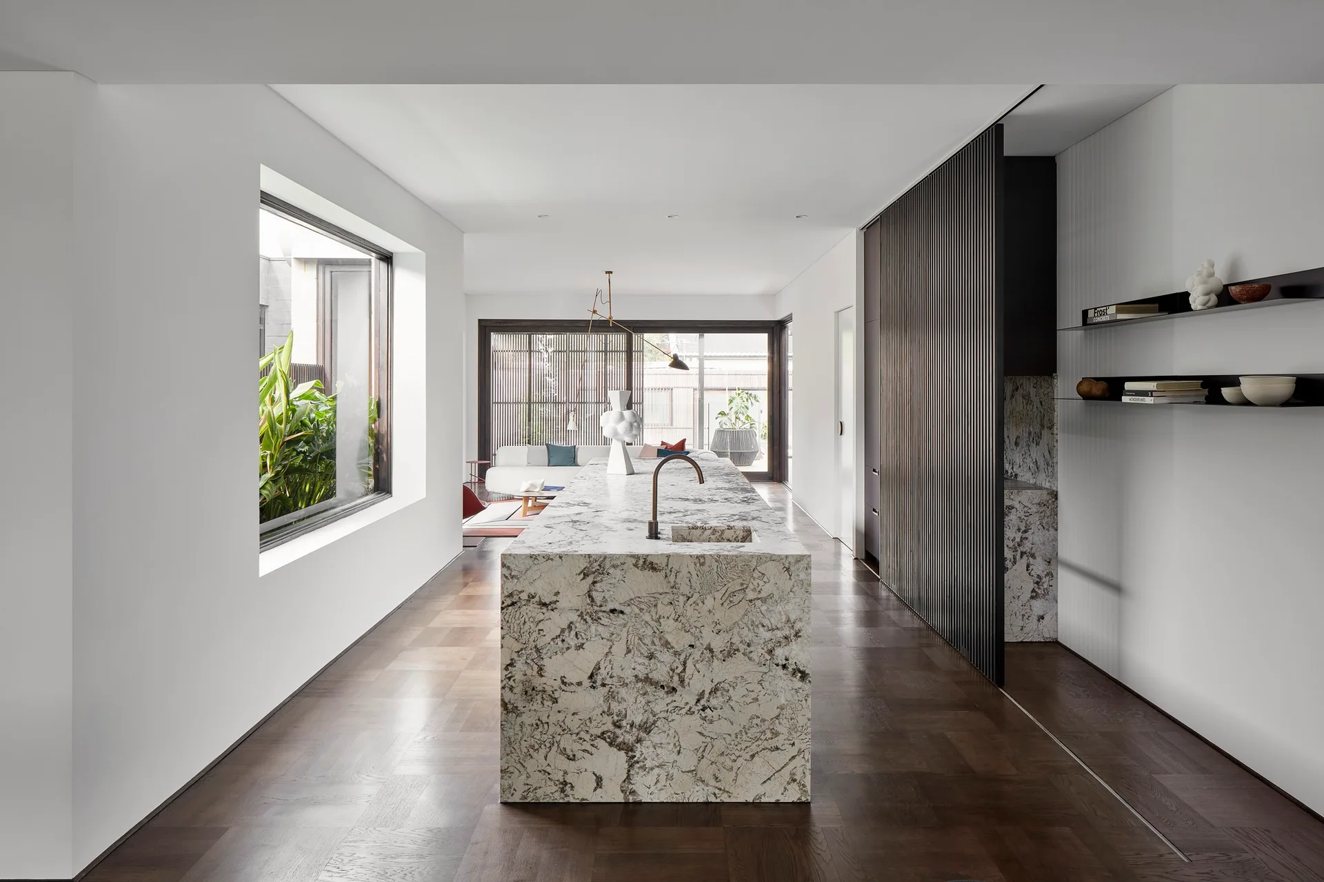 A minimalist kitchen features a large veined stone island, dark wood parquet flooring, a slatted wall partition, and a large window.