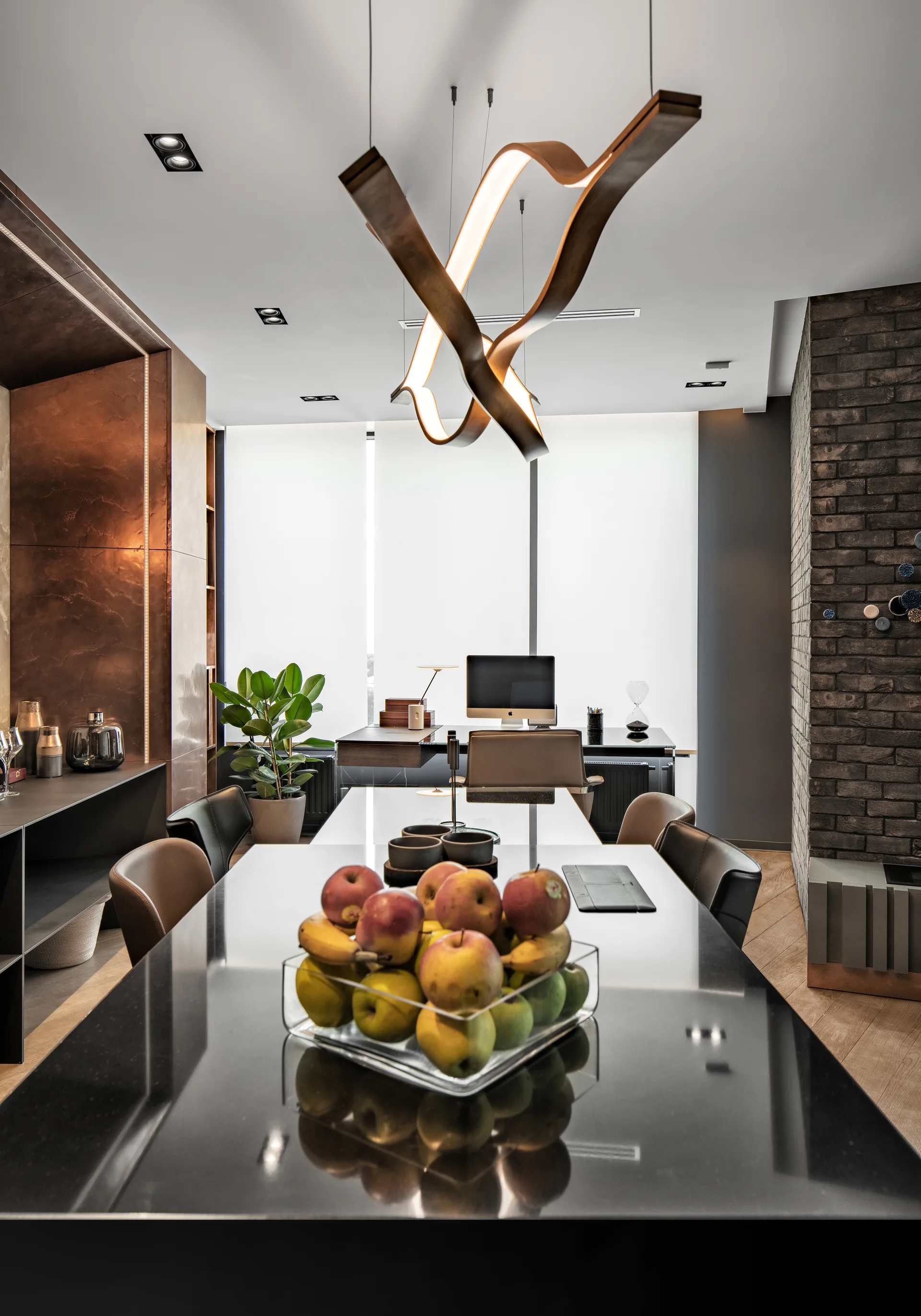 A modern workspace features a glossy black table, a curved wooden pendant light, and a desk in front of white roller blinds.