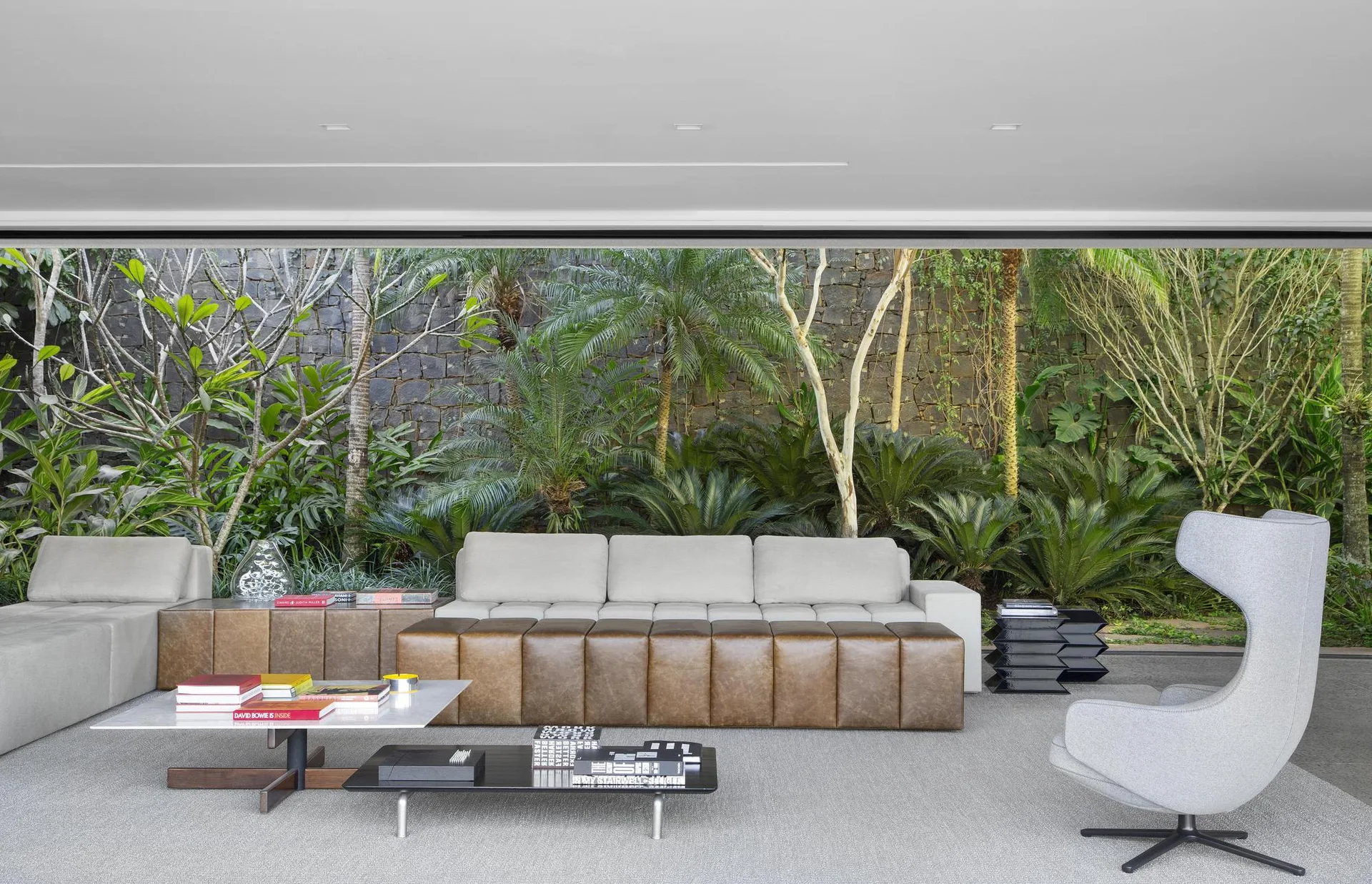 A modern living room features a grey sofa, a long brown leather bench, and a high-back swivel chair facing a glass wall with views of a lush green garden.