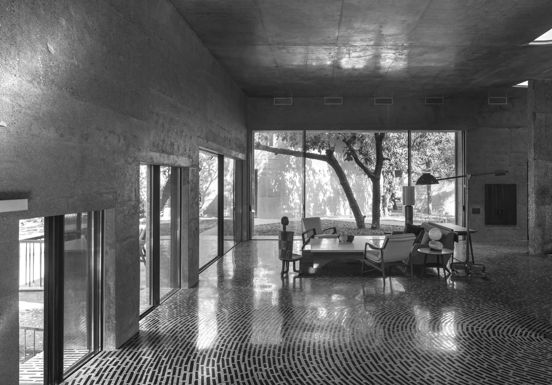 A spacious living area features rough concrete walls, mid-century modern furniture, and a polished floor with a radiating curved brick pattern.