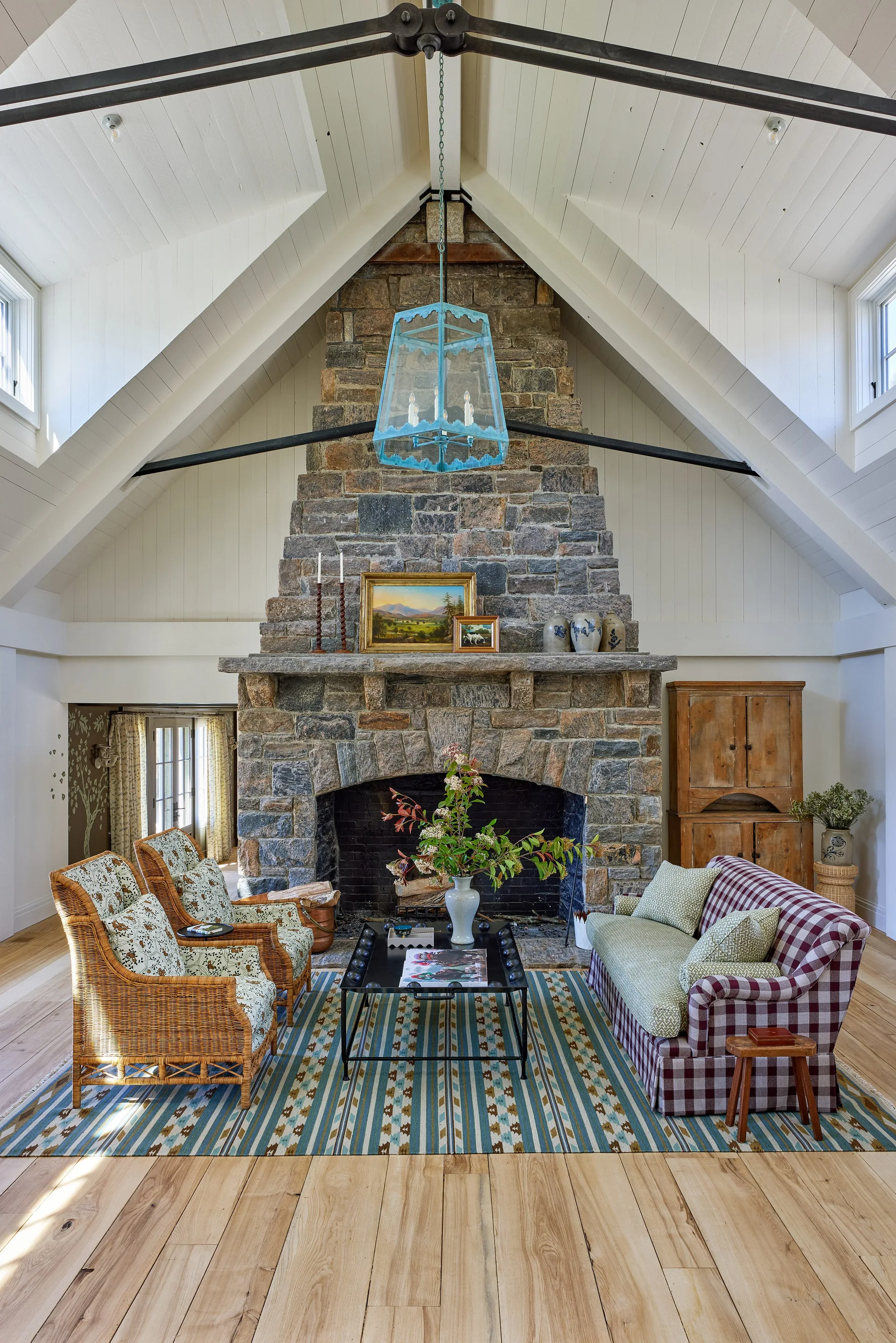 A vaulted living room features a tall stone fireplace, wicker armchairs, a checkered sofa, and a blue lantern pendant light.