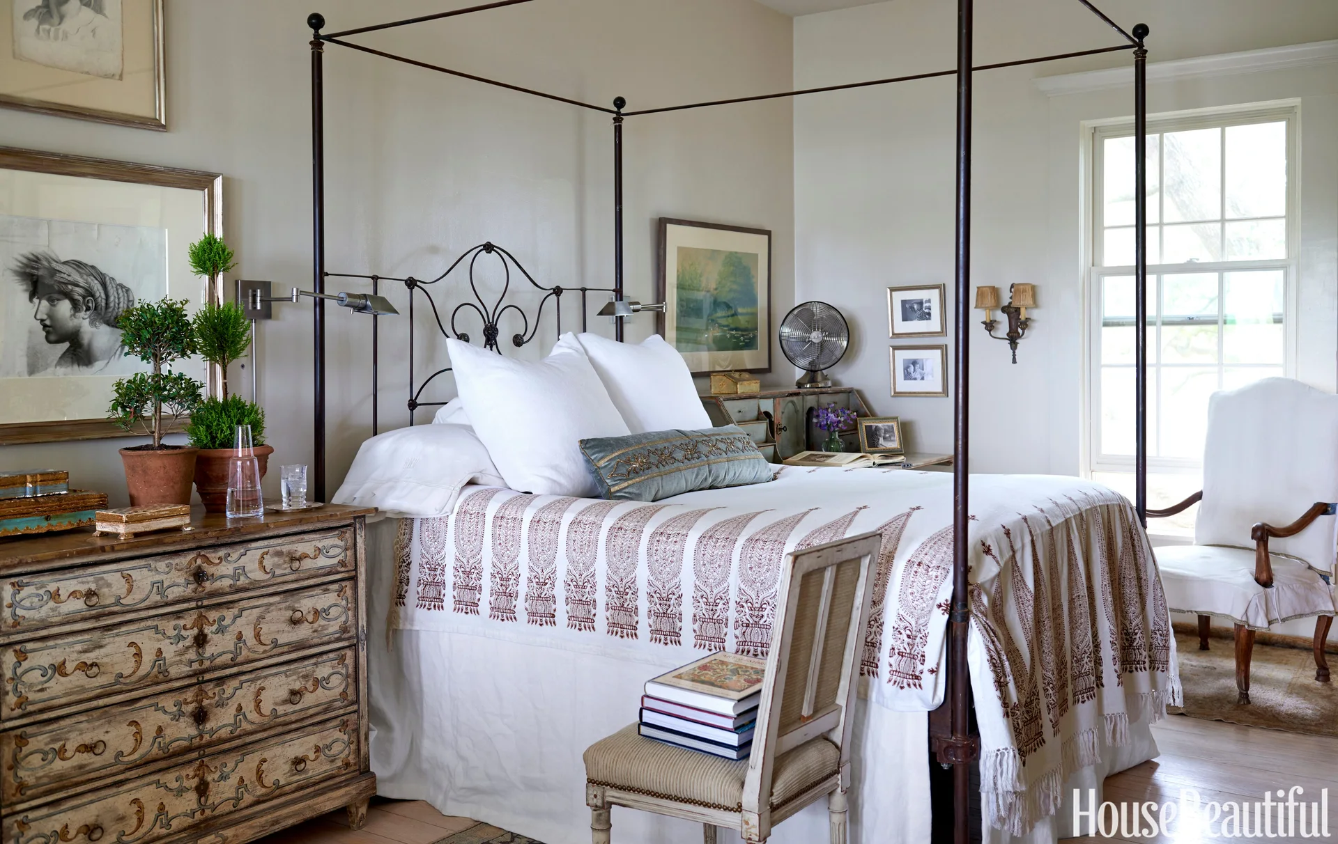 A traditional bedroom featuring a dark wrought iron four-poster bed, an ornate distressed wooden dresser, and beige walls.