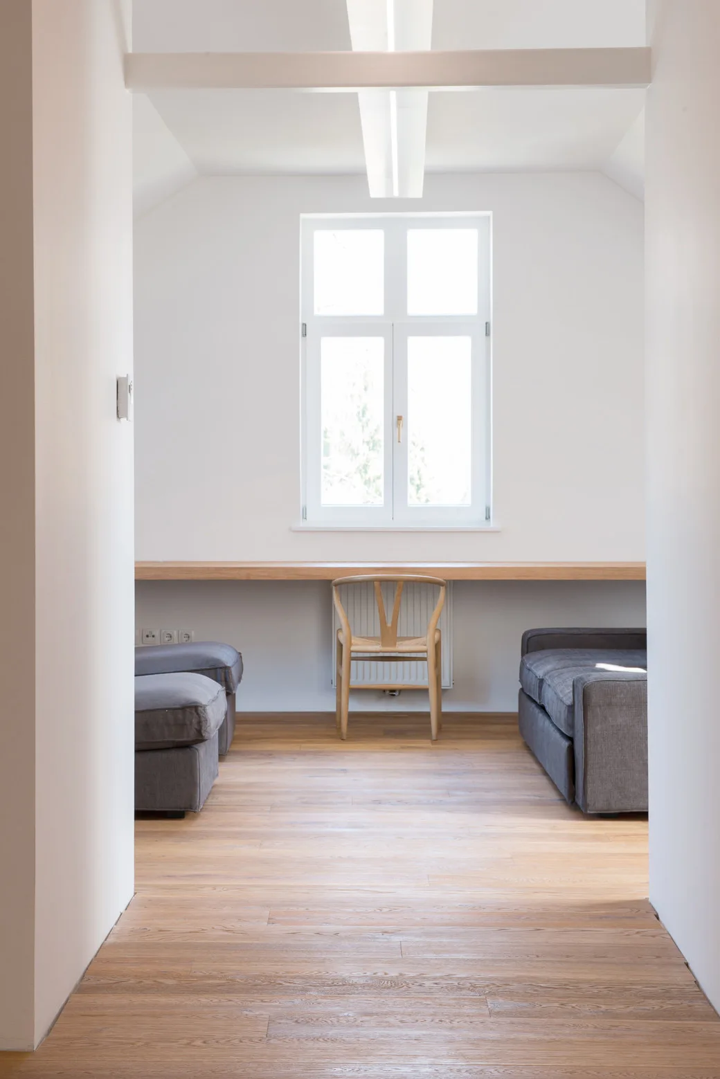 A minimalist workspace featuring a wall-to-wall wooden desk, a wishbone chair under a central window, and two symmetrical gray sofas.