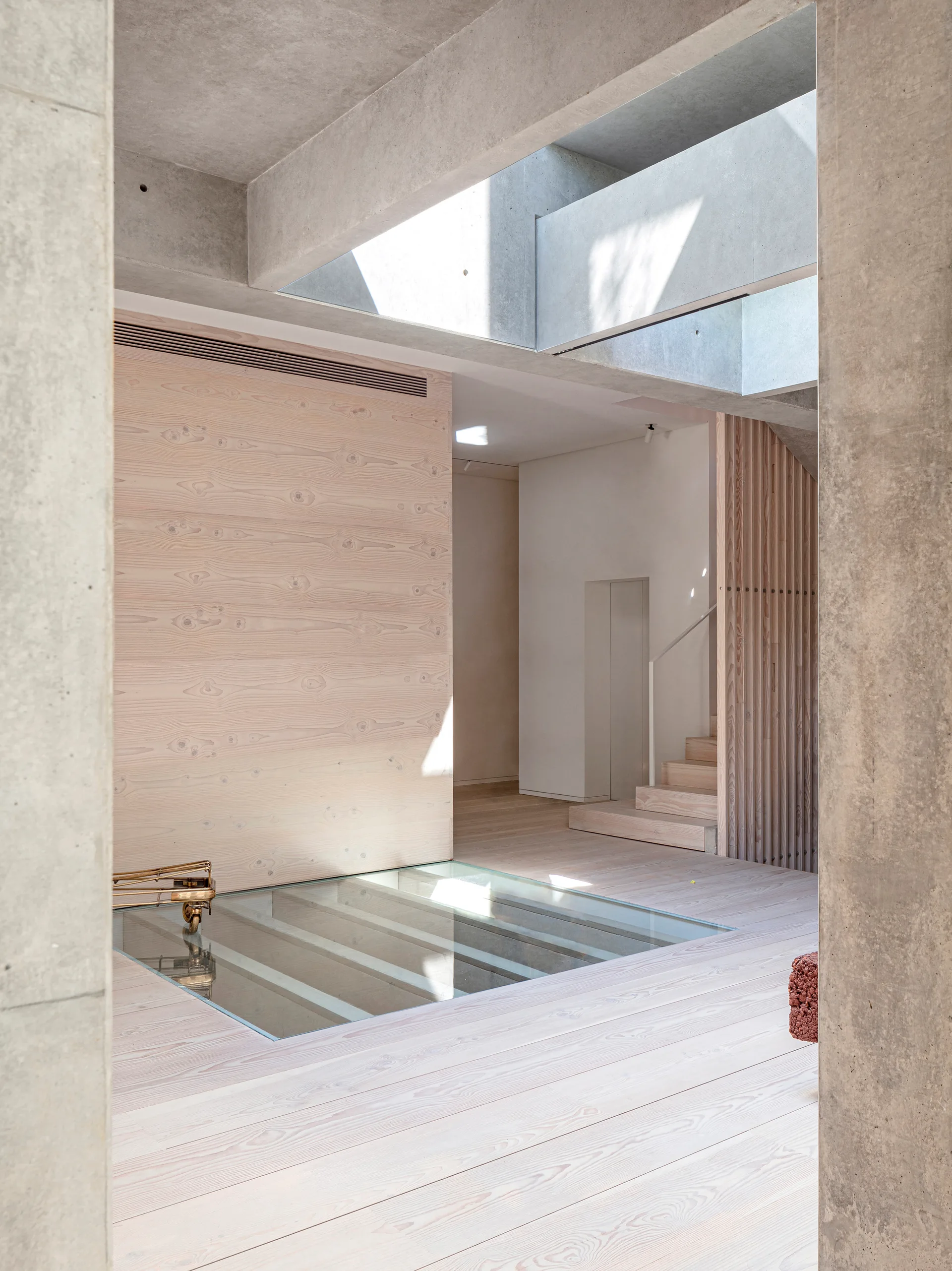 A minimalist modern interior with a pale wood floor, raw concrete ceiling beams with skylights, wood slat walls, and a glass floor insert.