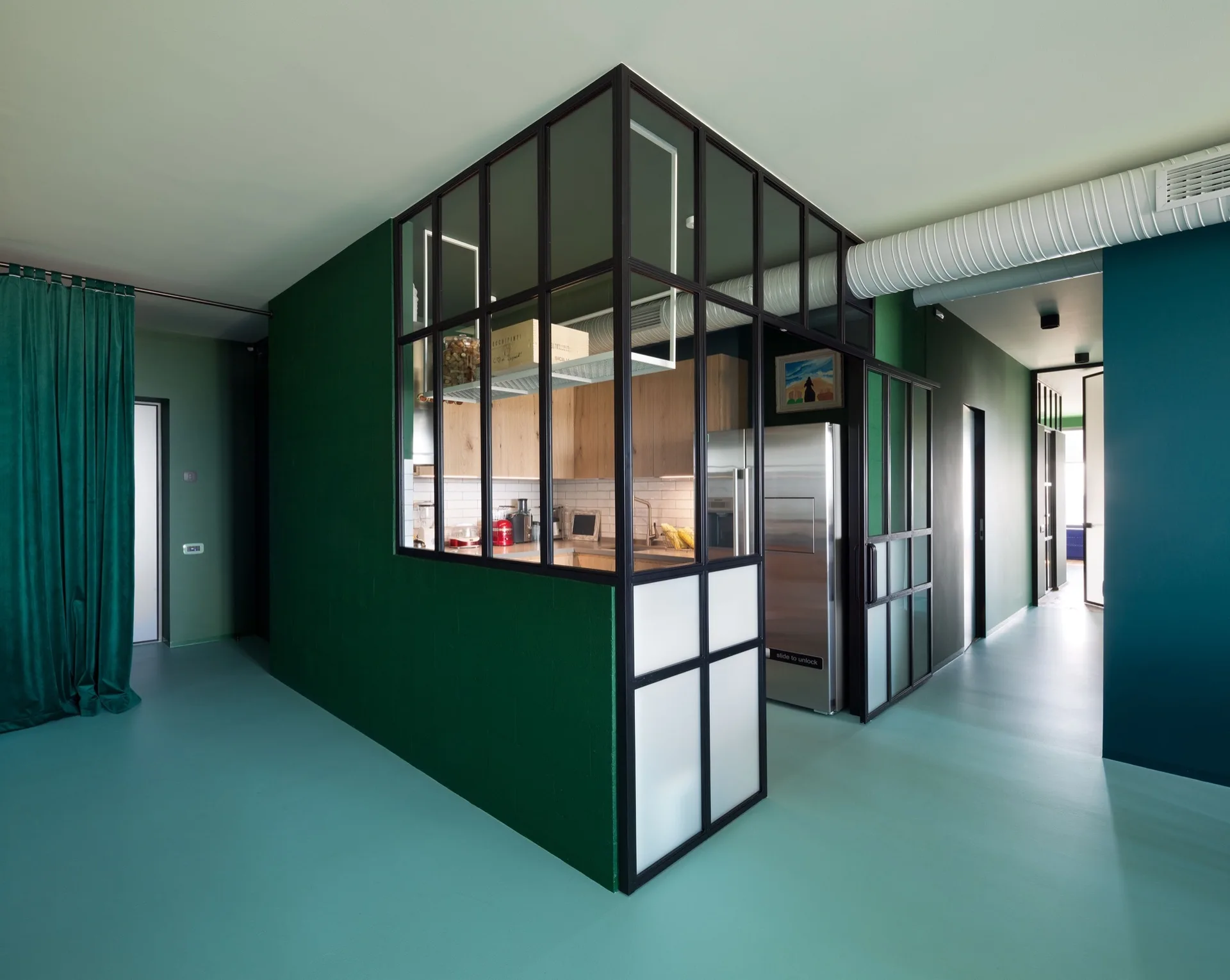 A kitchen with wooden cabinets is enclosed by a black metal-framed glass partition within a room with deep green walls and teal floors.