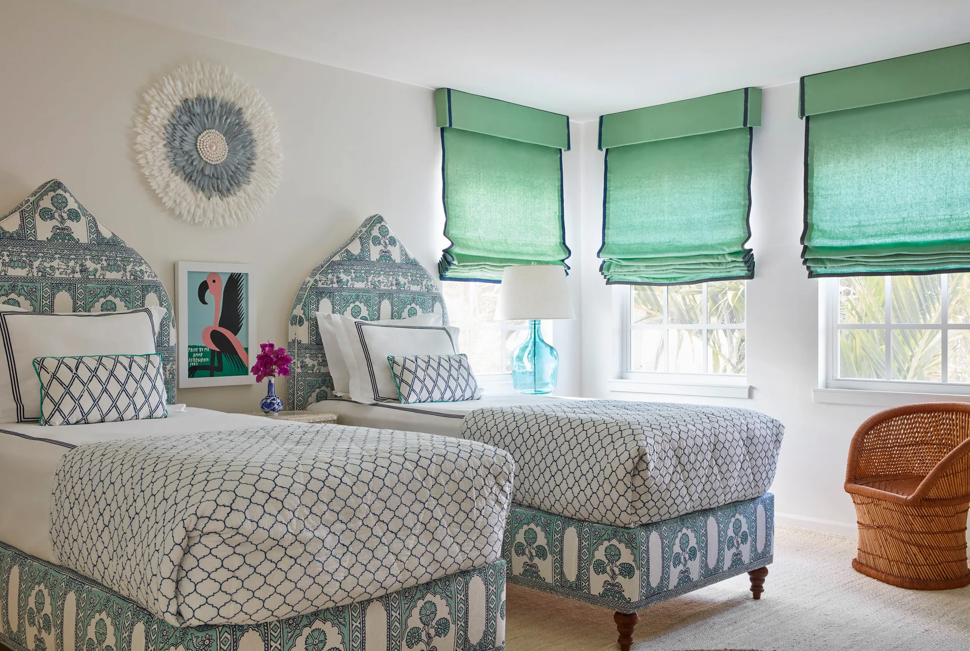 A twin bedroom features matching beds with teal patterned headboards, quilted coverlets, green fabric roman shades, and a woven rattan chair in the corner.