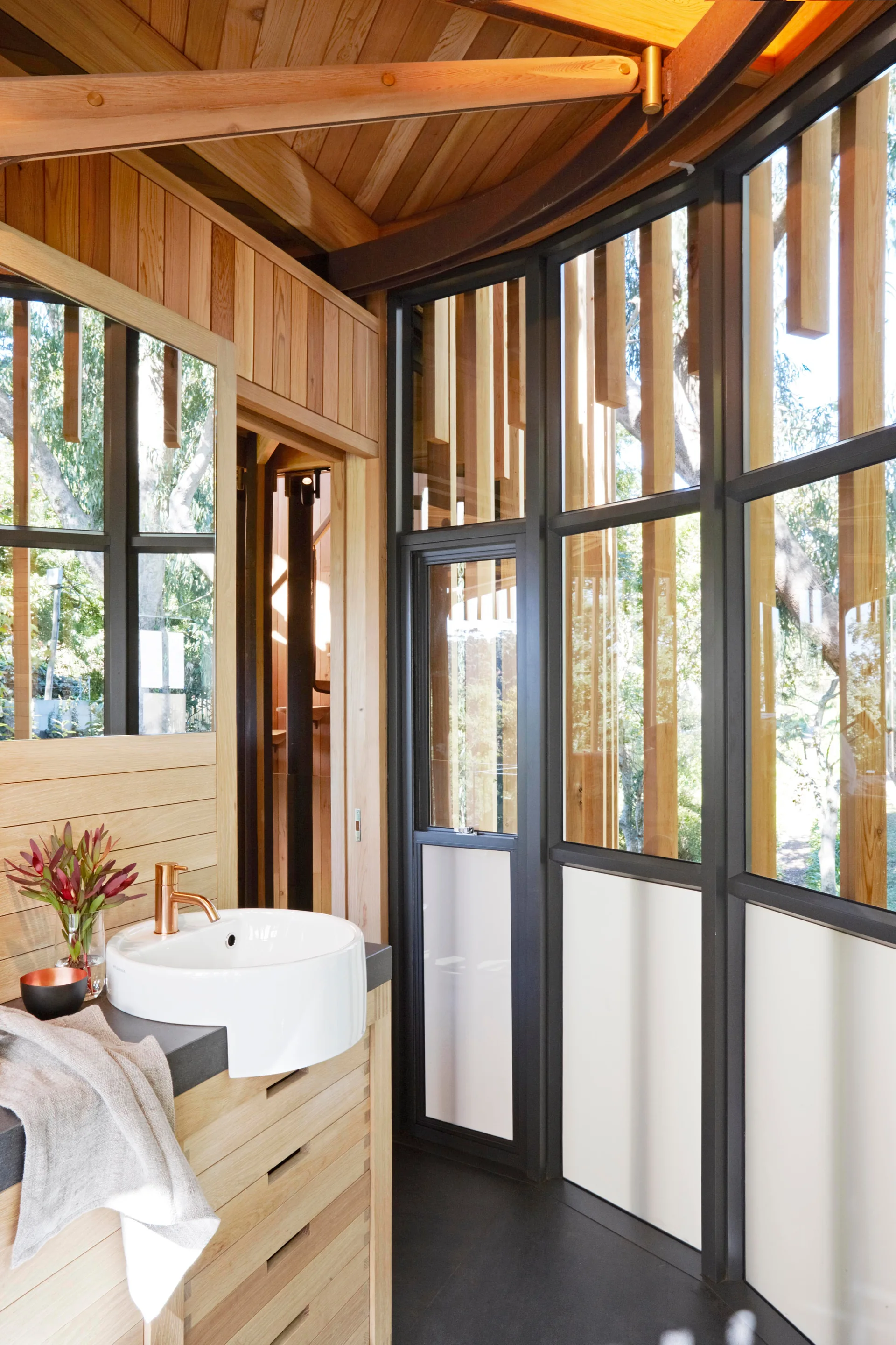 A modern bathroom features a wooden vanity with a white vessel sink, vertical wood-paneled walls, and floor-to-ceiling windows with dark frames.