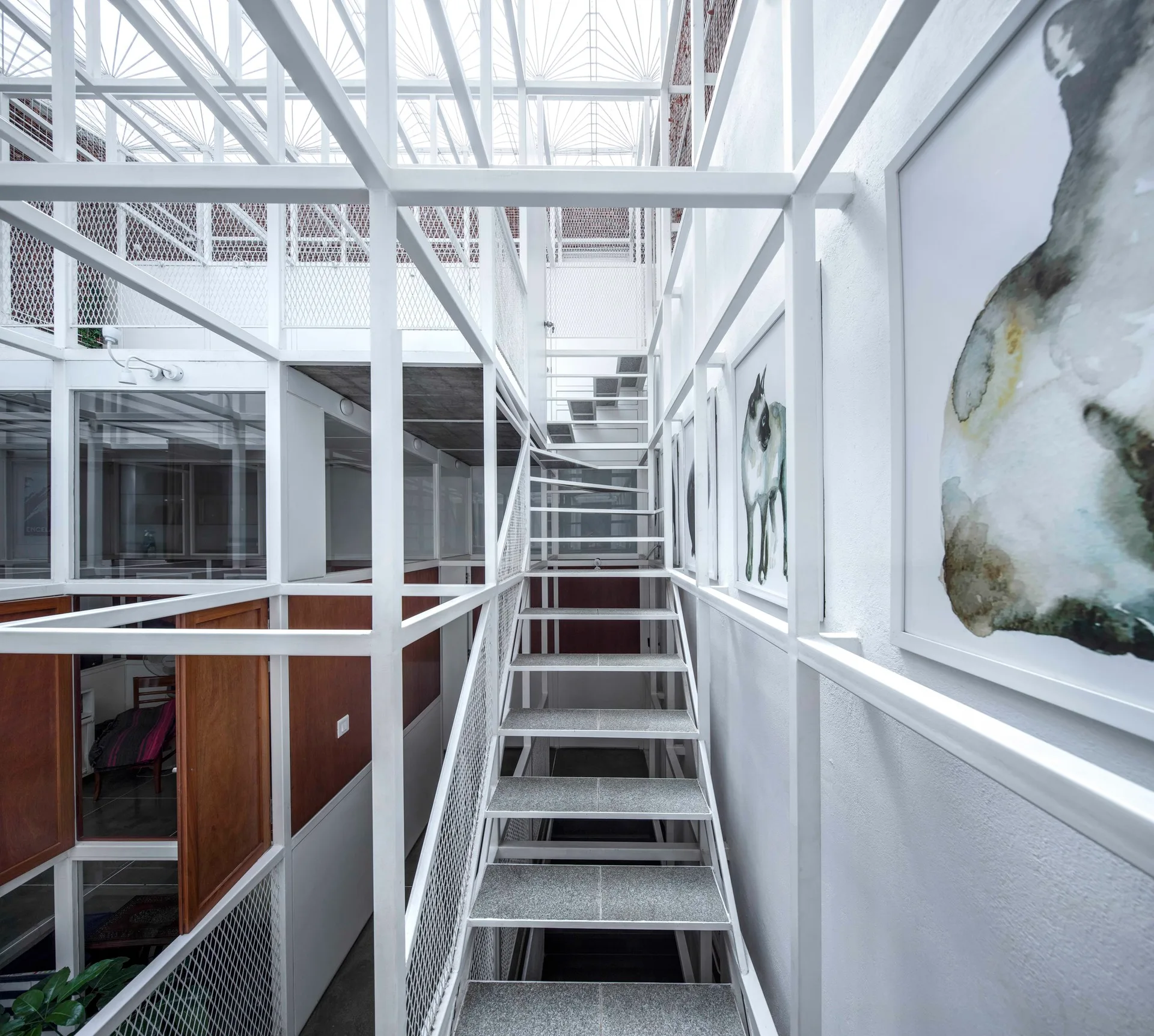 A modern staircase with open risers ascends through a white metal grid structure, flanked by wood-paneled walls and framed art.