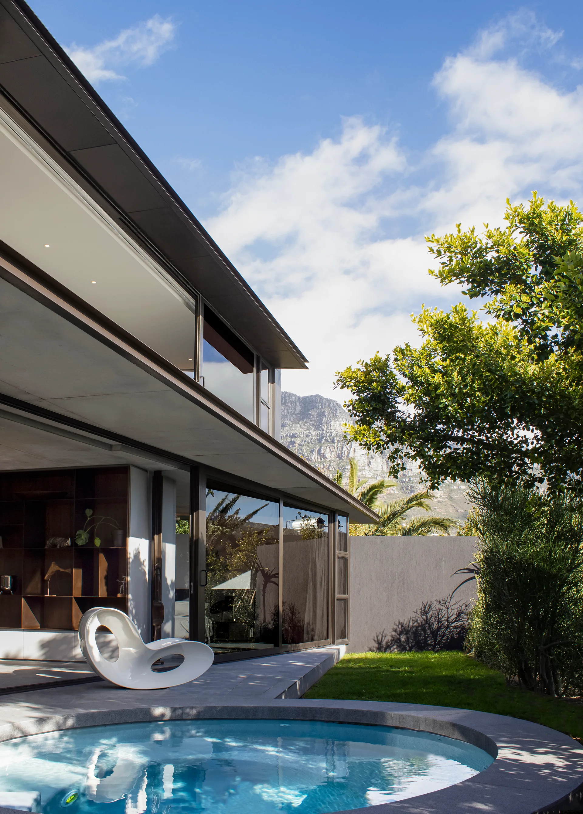 A circular blue pool rests on a grey stone patio beside a modern home with large glass sliding doors and a white sculptural chair.
