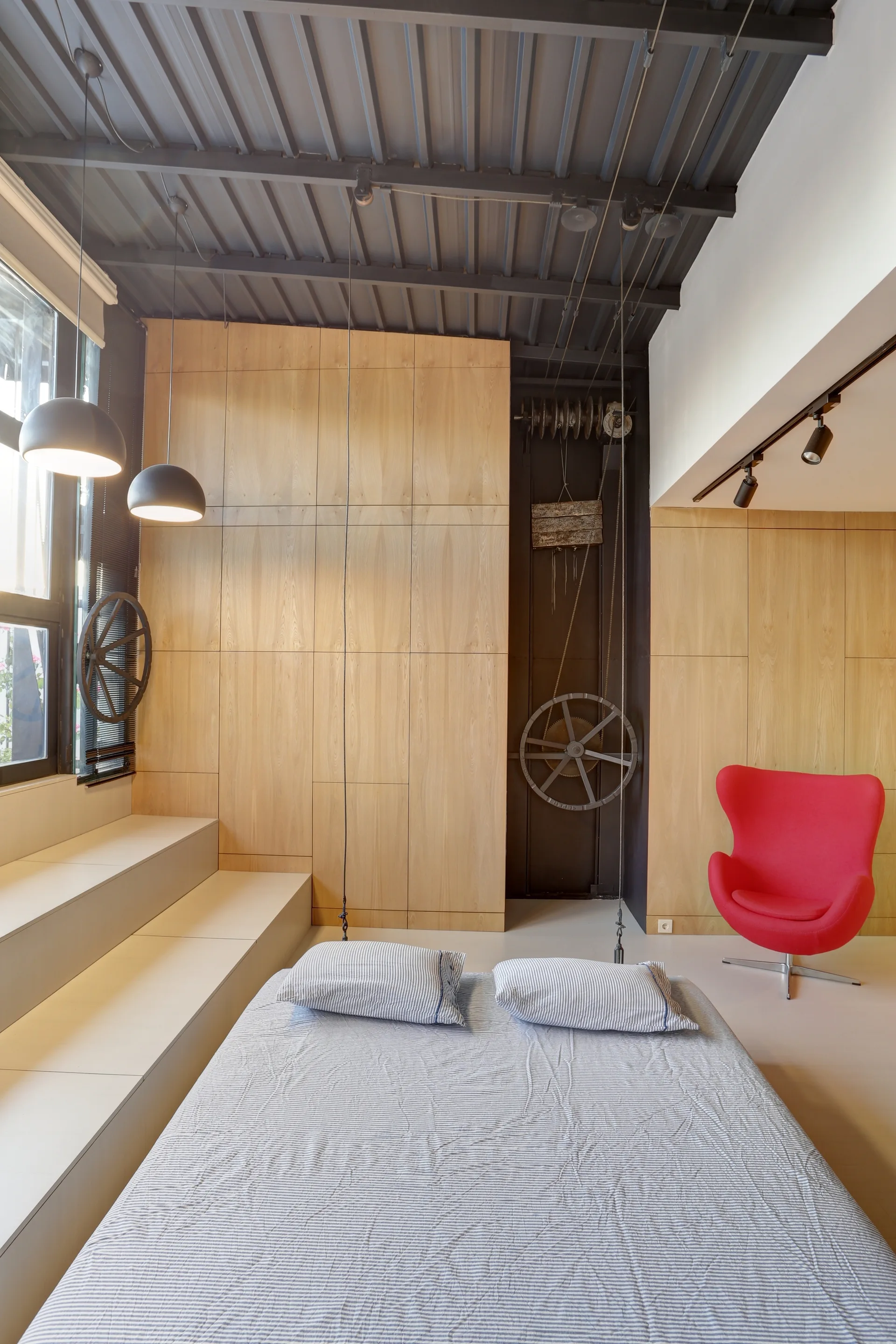 An industrial bedroom featuring light wood wall panels, a dark corrugated metal ceiling, a stepped platform near a window, and a red armchair.