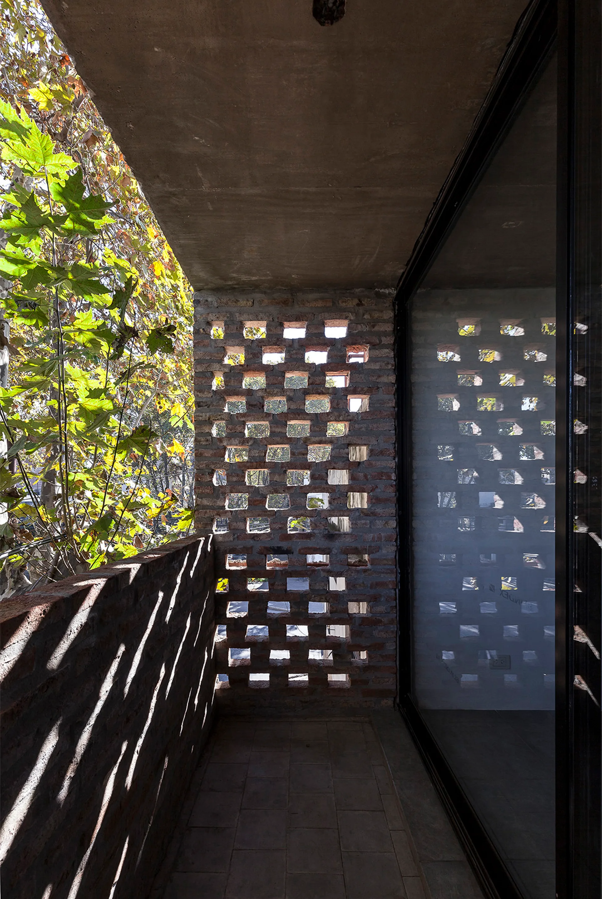 A modern outdoor balcony featuring a perforated red brick wall screen, an exposed concrete ceiling, and a sliding glass door.
