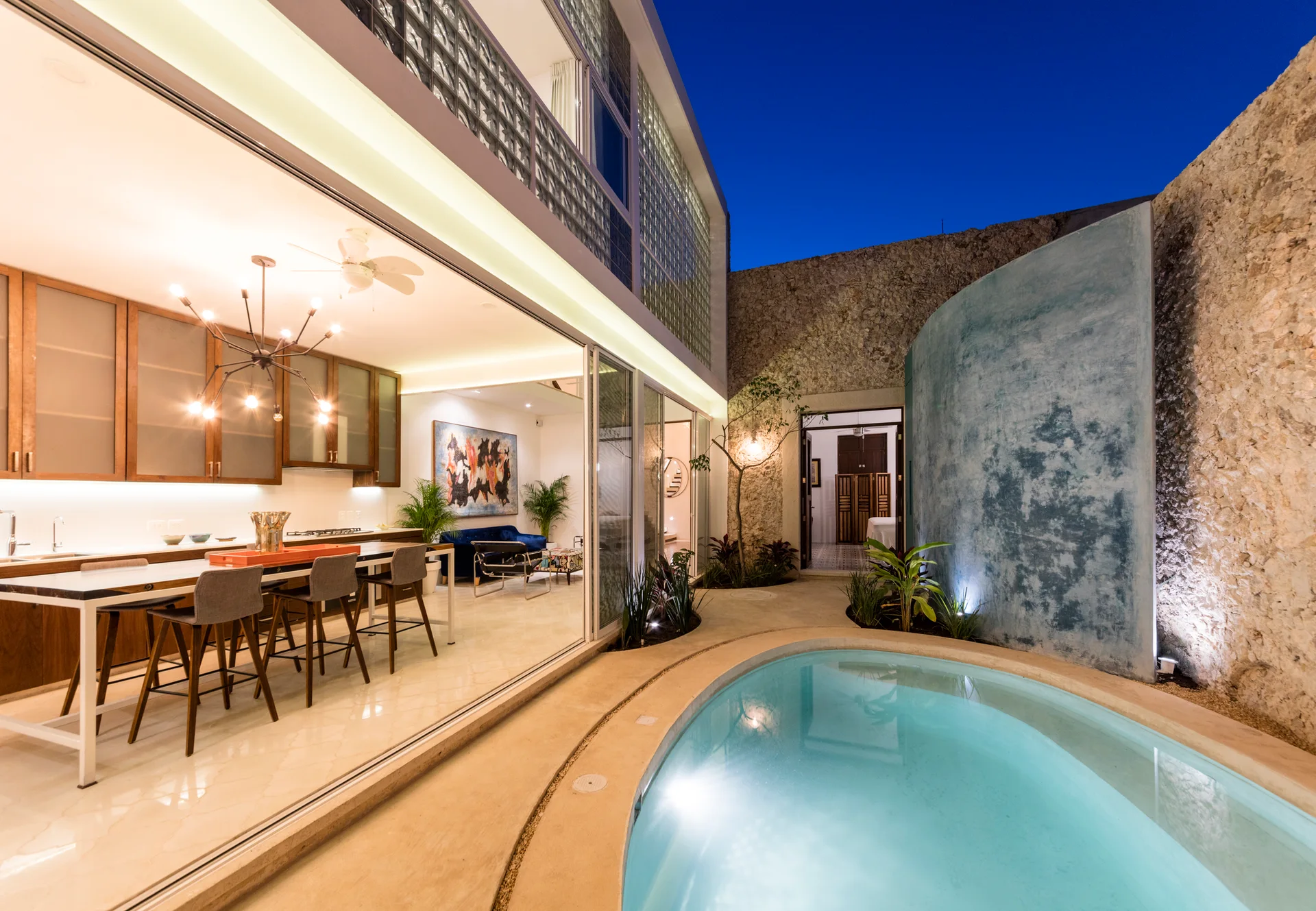 A modern kitchen with wood cabinets opens through sliding glass doors to a courtyard patio with a curved pool and rustic stone walls at night.