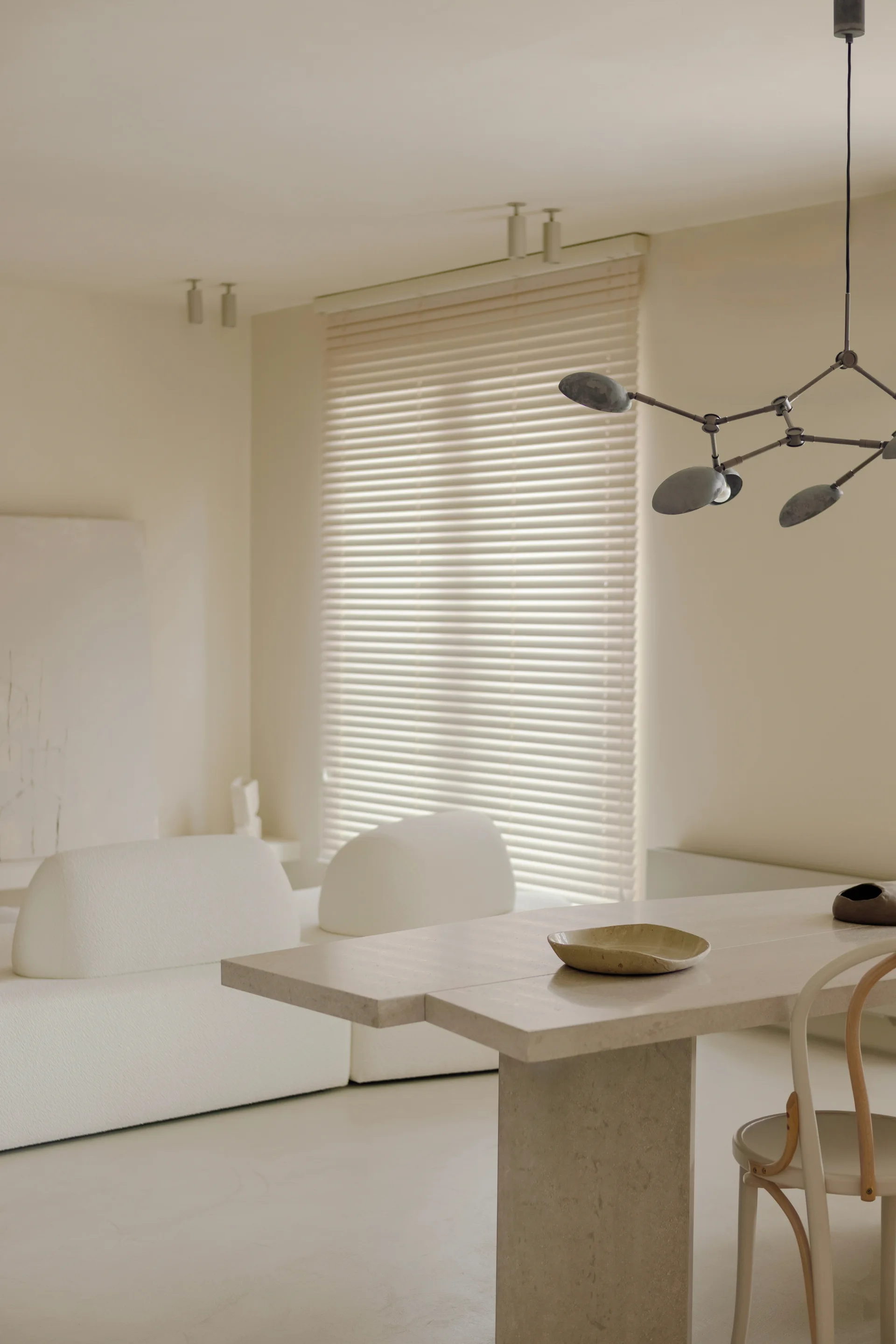 A minimalist cream dining room with a thick stone table, a curved light wood chair, a branching dark pendant, and horizontal window blinds.