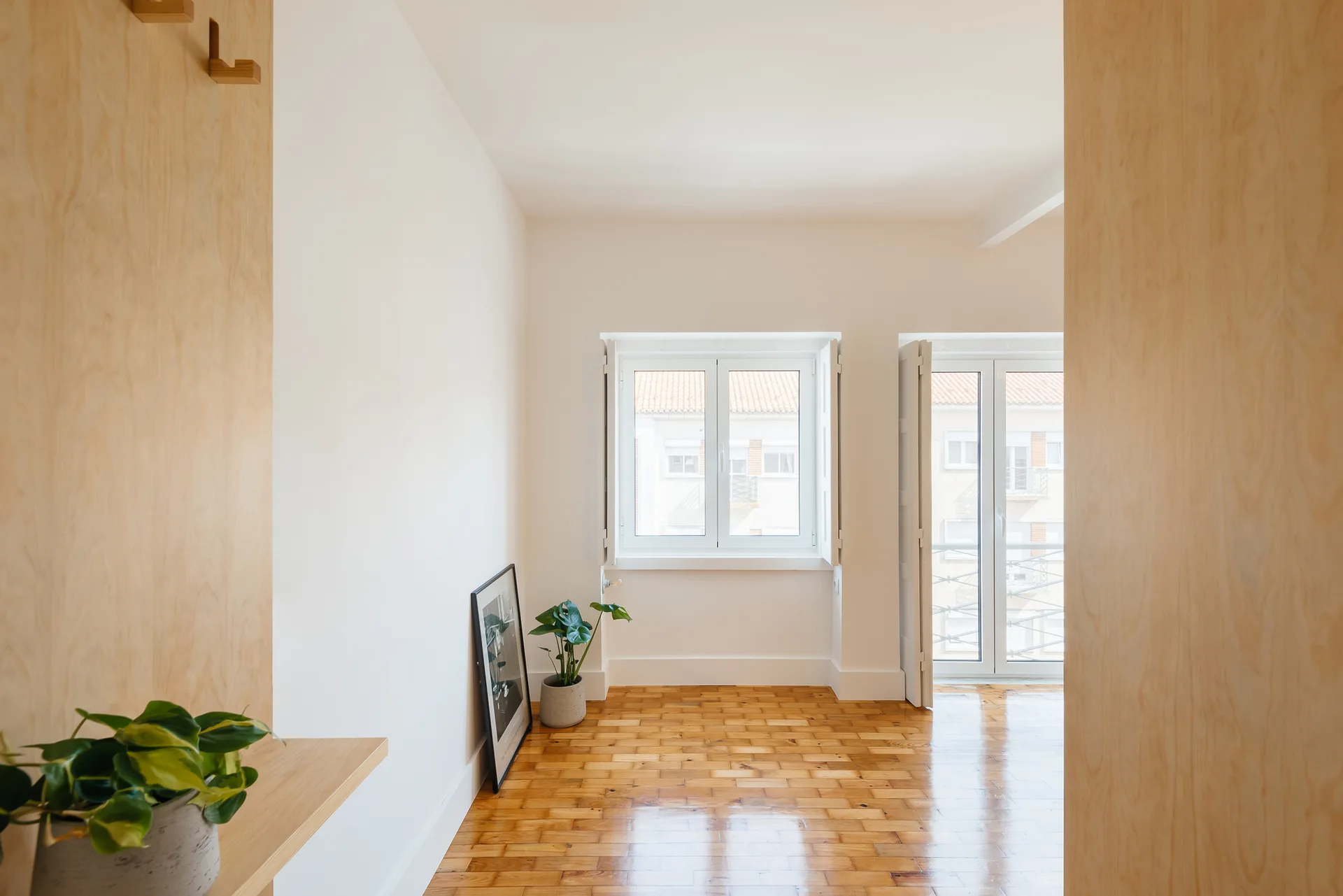 A bright empty room featuring polished wood parquet floors, white walls with interior shutters, and light wood wall panels framing the entrance.